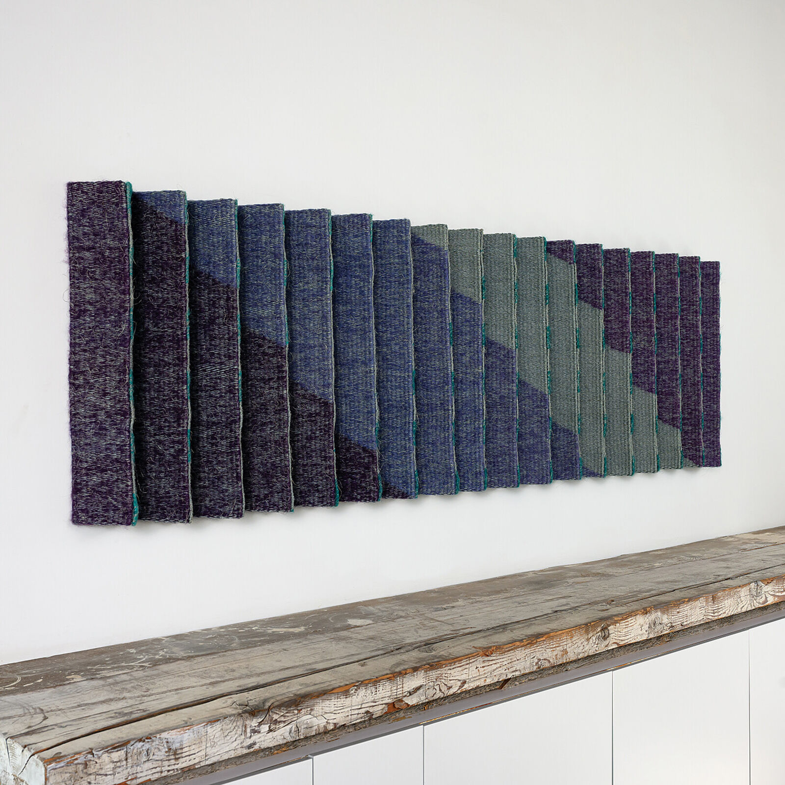 A geometric textile wall hanging featuring a pleated, accordion-fold design in teal, purple, and grey tones.