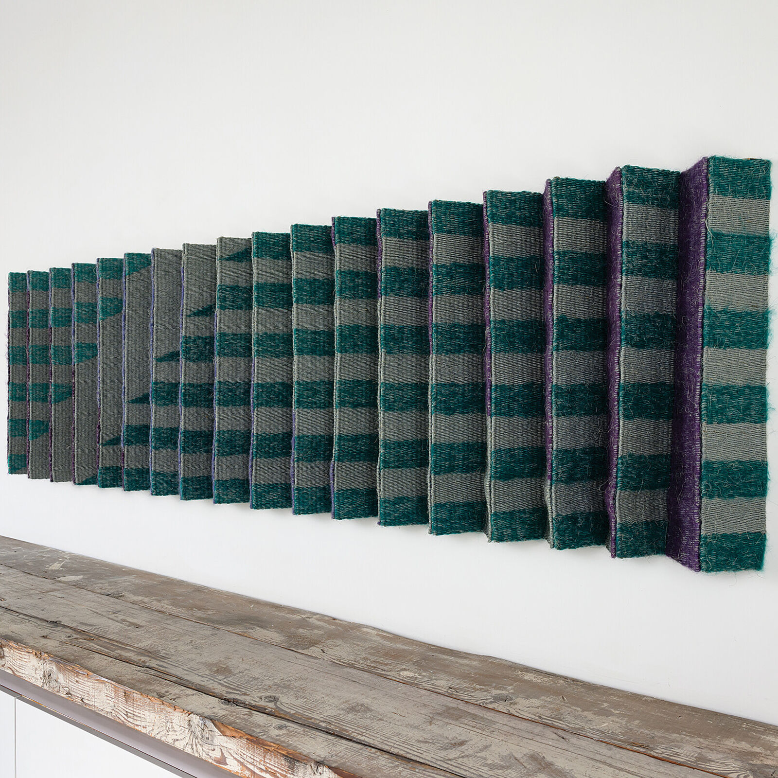 A geometric textile wall hanging featuring a pleated, accordion-fold design in teal, purple, and grey tones.