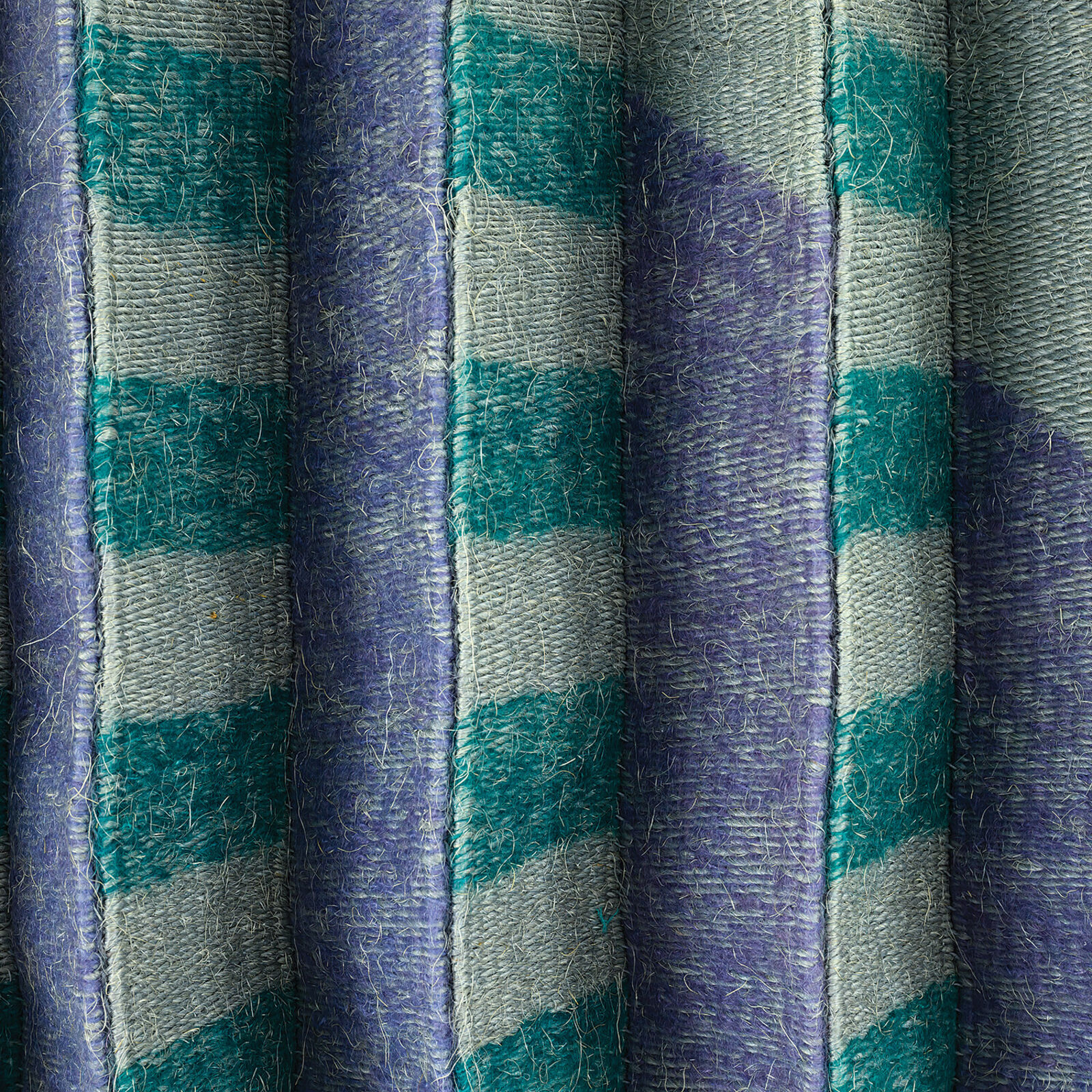 A geometric textile wall hanging featuring a pleated, accordion-fold design in teal, purple, and grey tones.