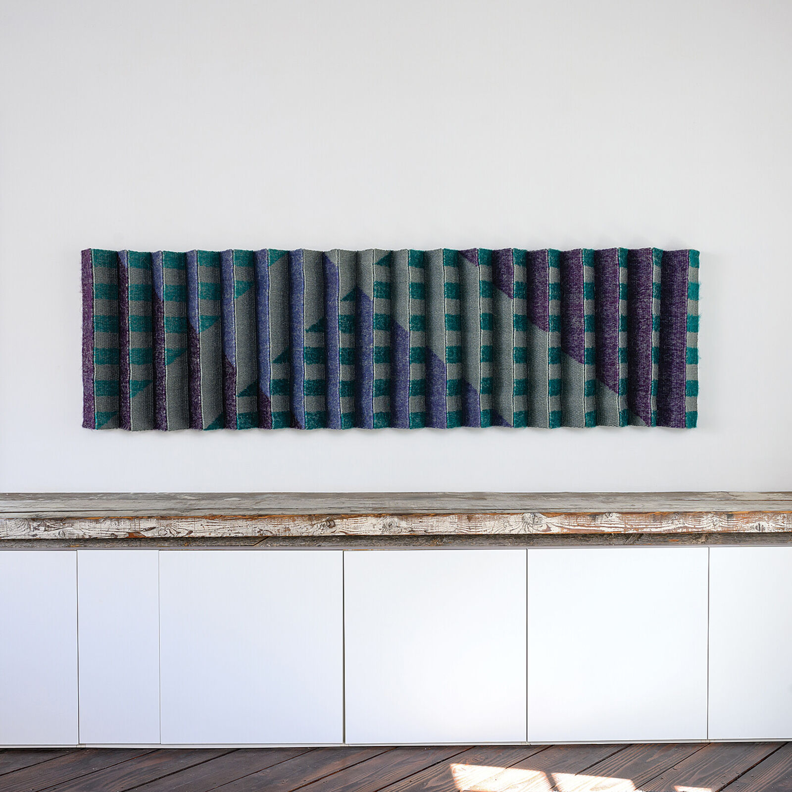 A geometric textile wall hanging featuring a pleated, accordion-fold design in teal, purple, and grey tones.