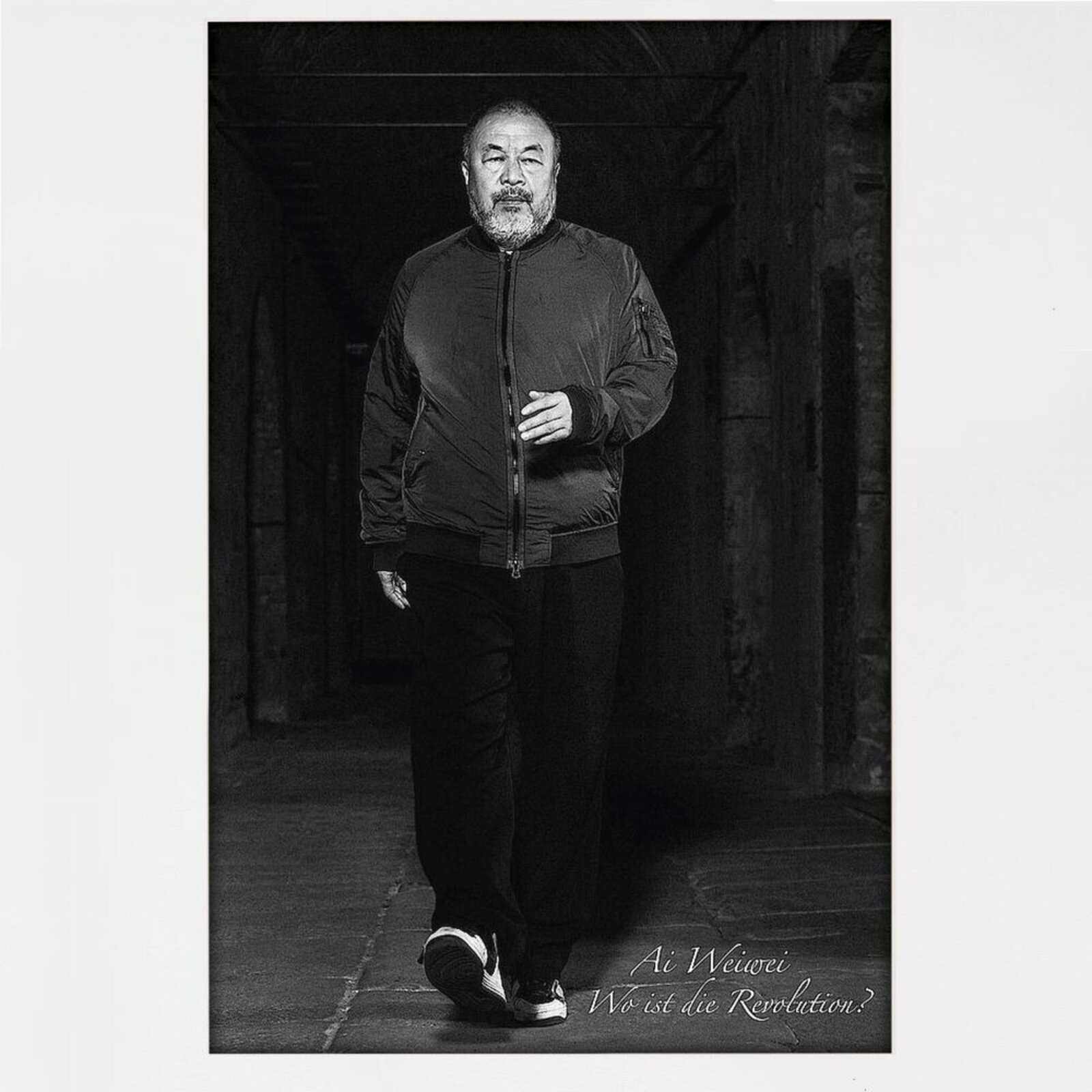 A black-and-white portrait of a bearded man walking forward in a dimly lit corridor, wearing a bomber jacket and sneakers.