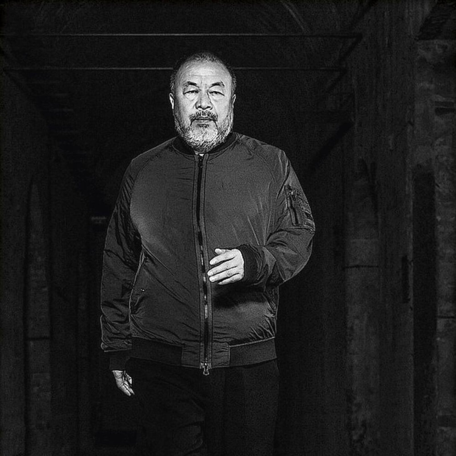 A black-and-white portrait of a bearded man walking forward in a dimly lit corridor, wearing a bomber jacket and sneakers.