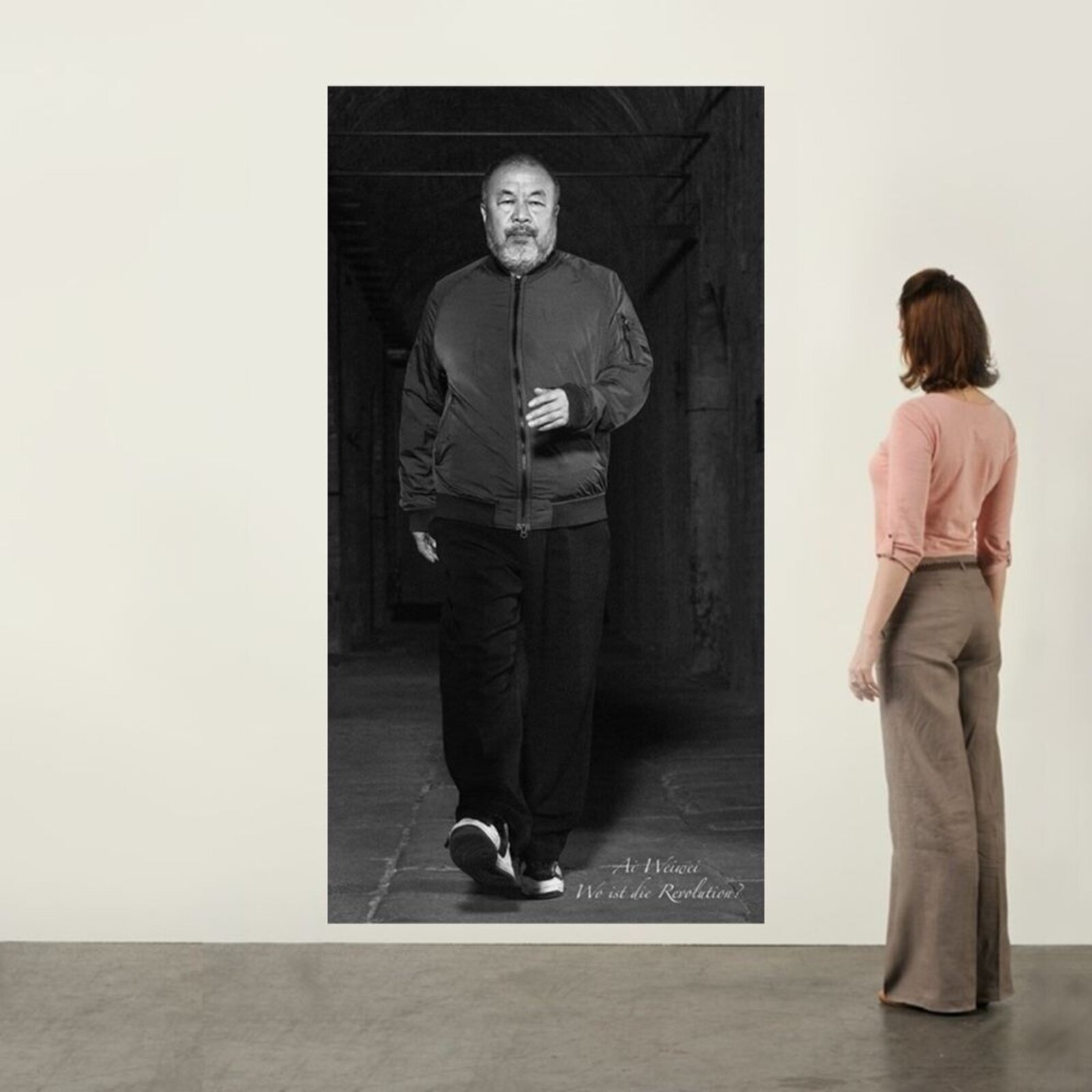A black-and-white portrait of a bearded man walking forward in a dimly lit corridor, wearing a bomber jacket and sneakers.
