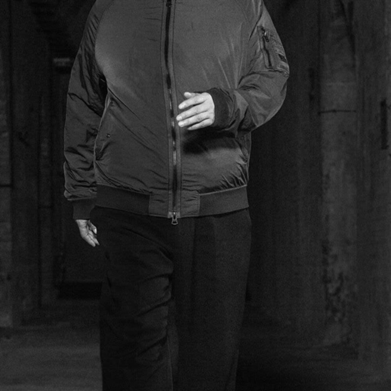 A black-and-white portrait of a bearded man walking forward in a dimly lit corridor, wearing a bomber jacket and sneakers.