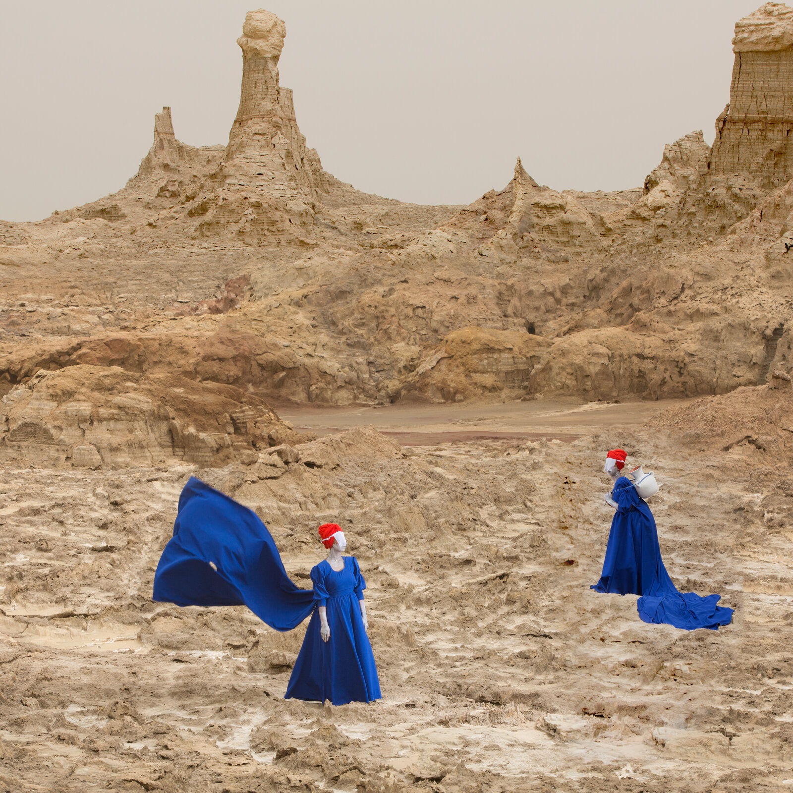 A surreal desert scene featuring two figures in blue gowns against eroded rock formations.