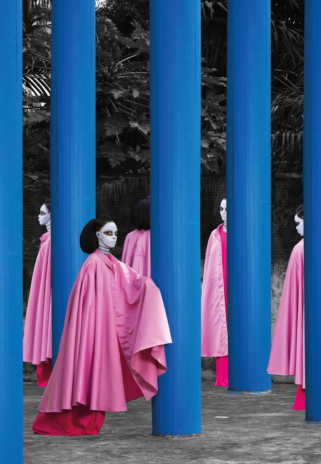 A surreal scene featuring figures in pink robes and white masks standing among tall blue pillars.