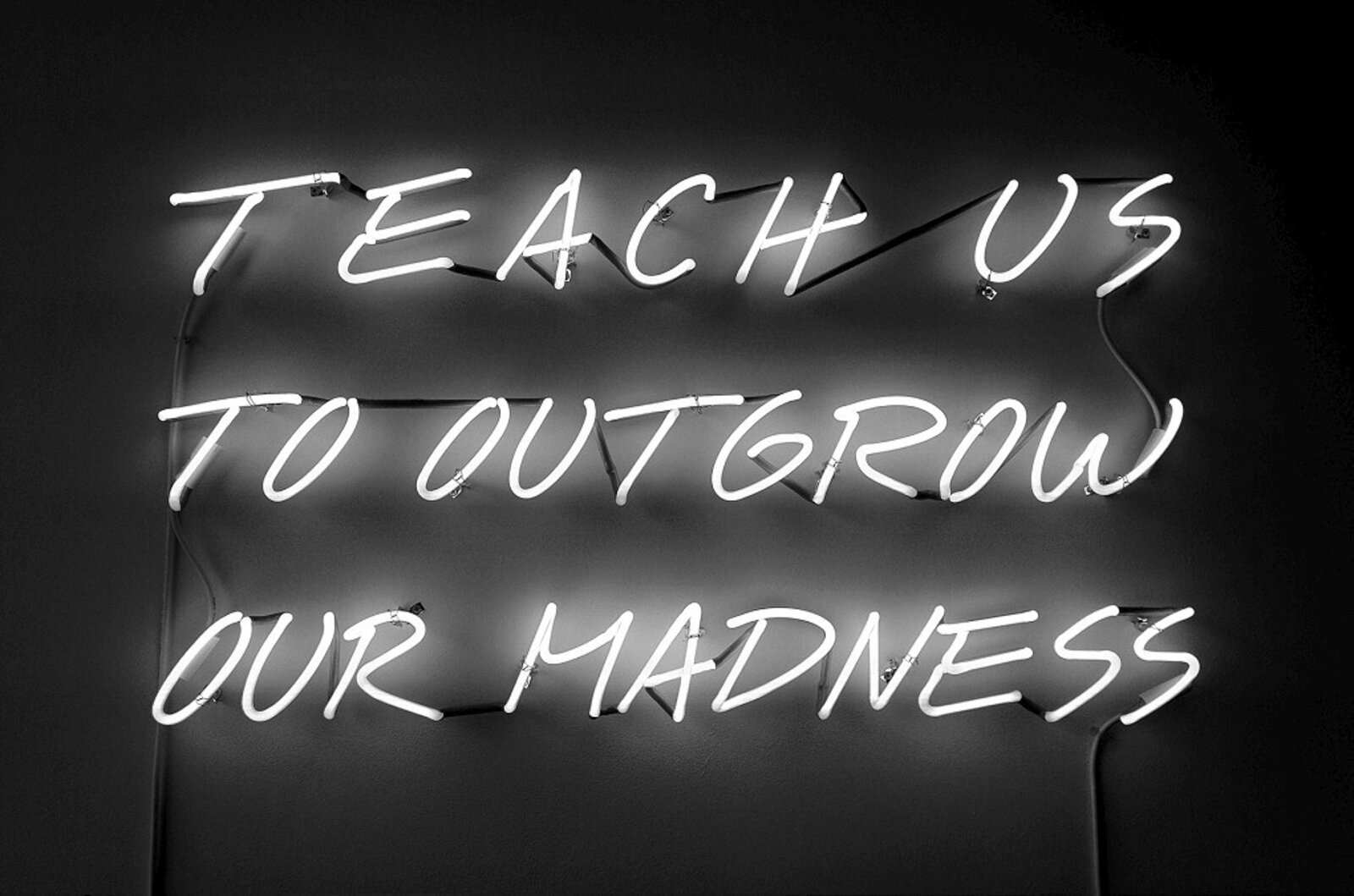 A black-and-white neon sign displaying the phrase 'TEACH US TO OUTGROW OUR MADNESS' in a handwritten style.