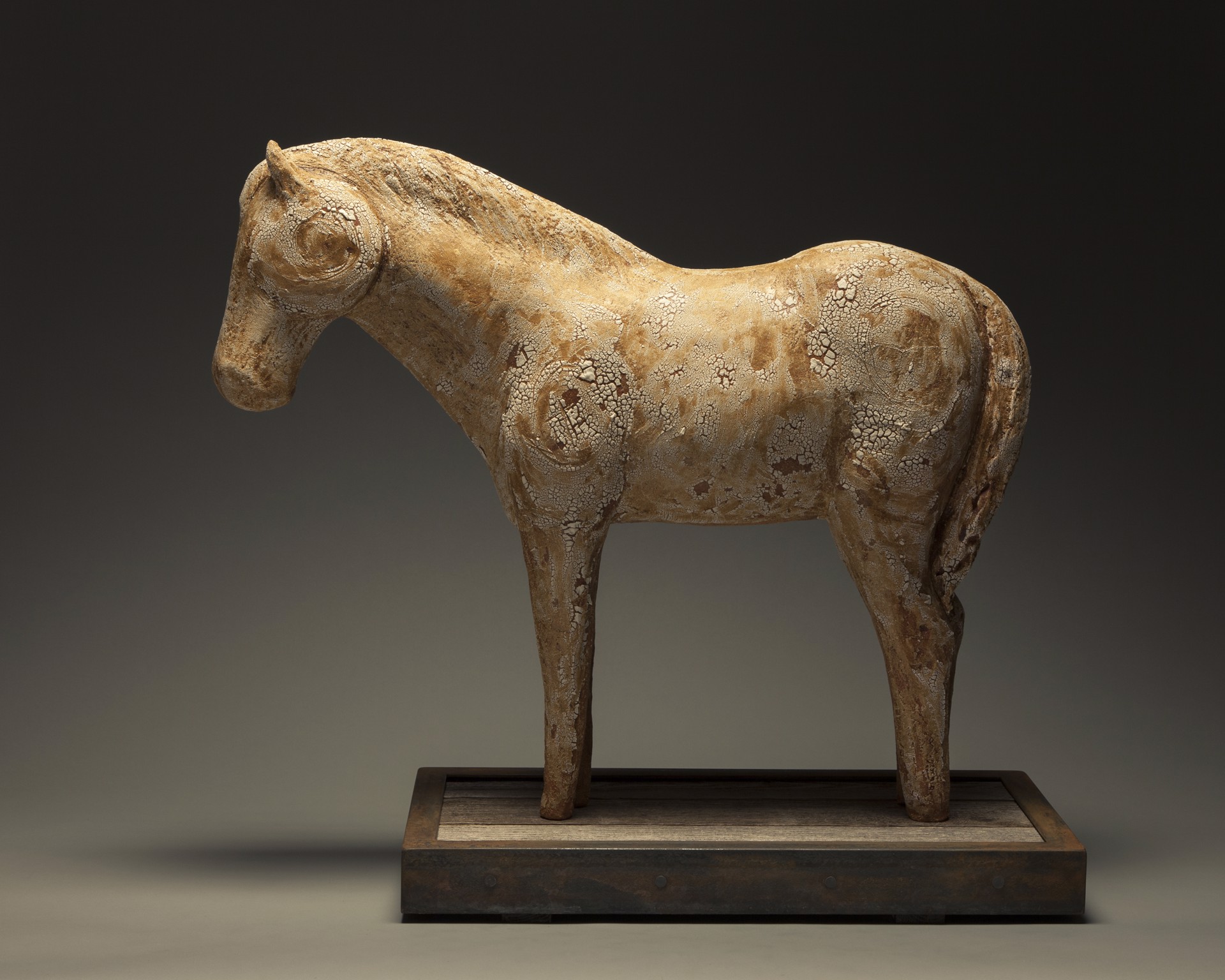A weathered horse sculpture stands on a dark wooden base against a neutral gradient background.