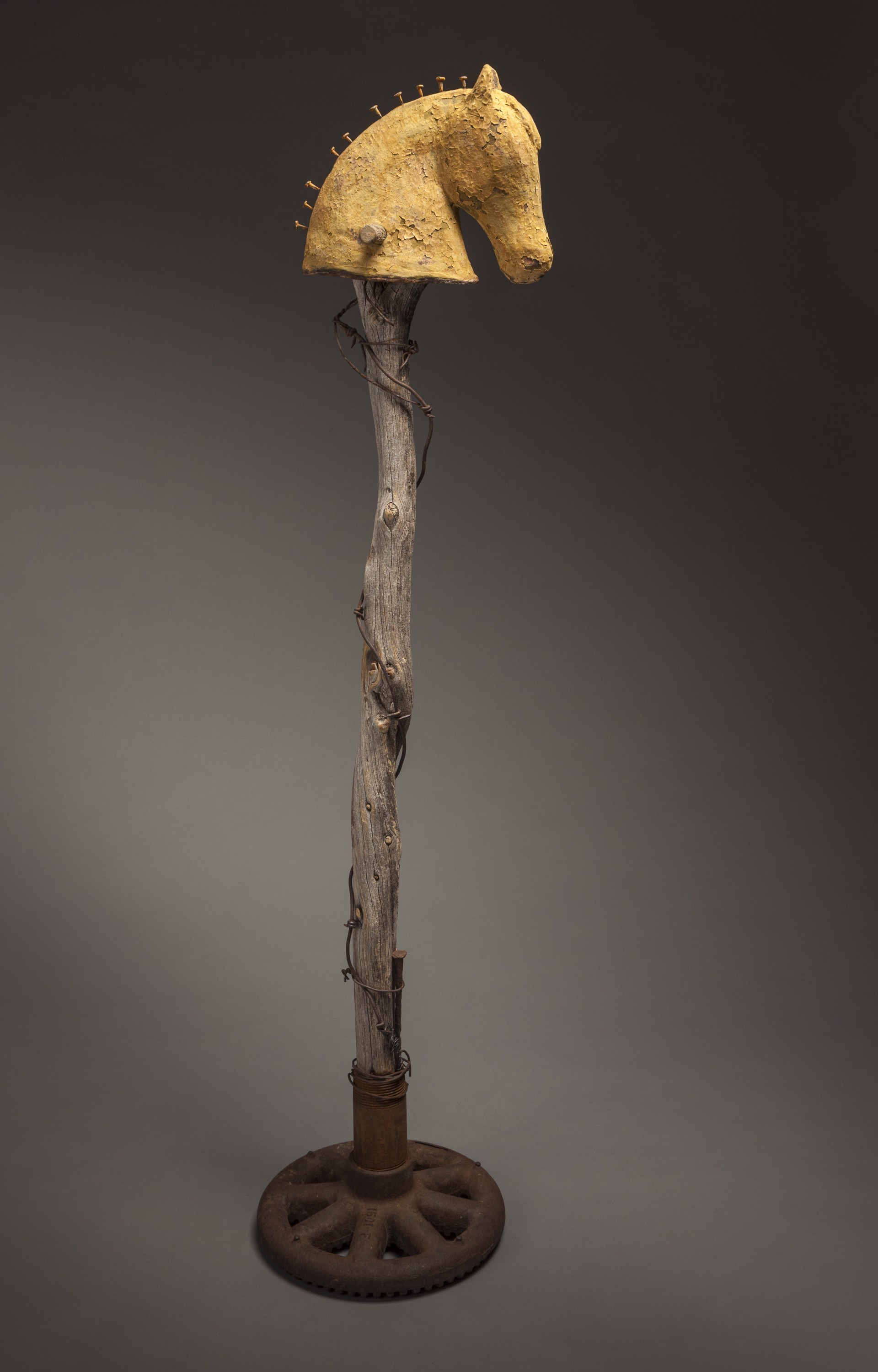 A whimsical sculpture featuring a horse head mounted on a rustic, vine-covered staff that rests on an industrial wheel base.