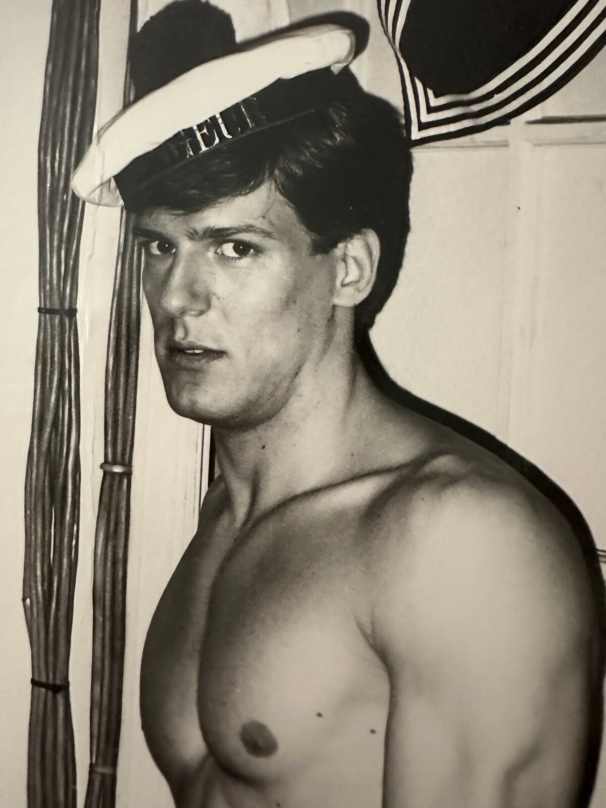 A shirtless man in a sailor's hat standing in front of a door.