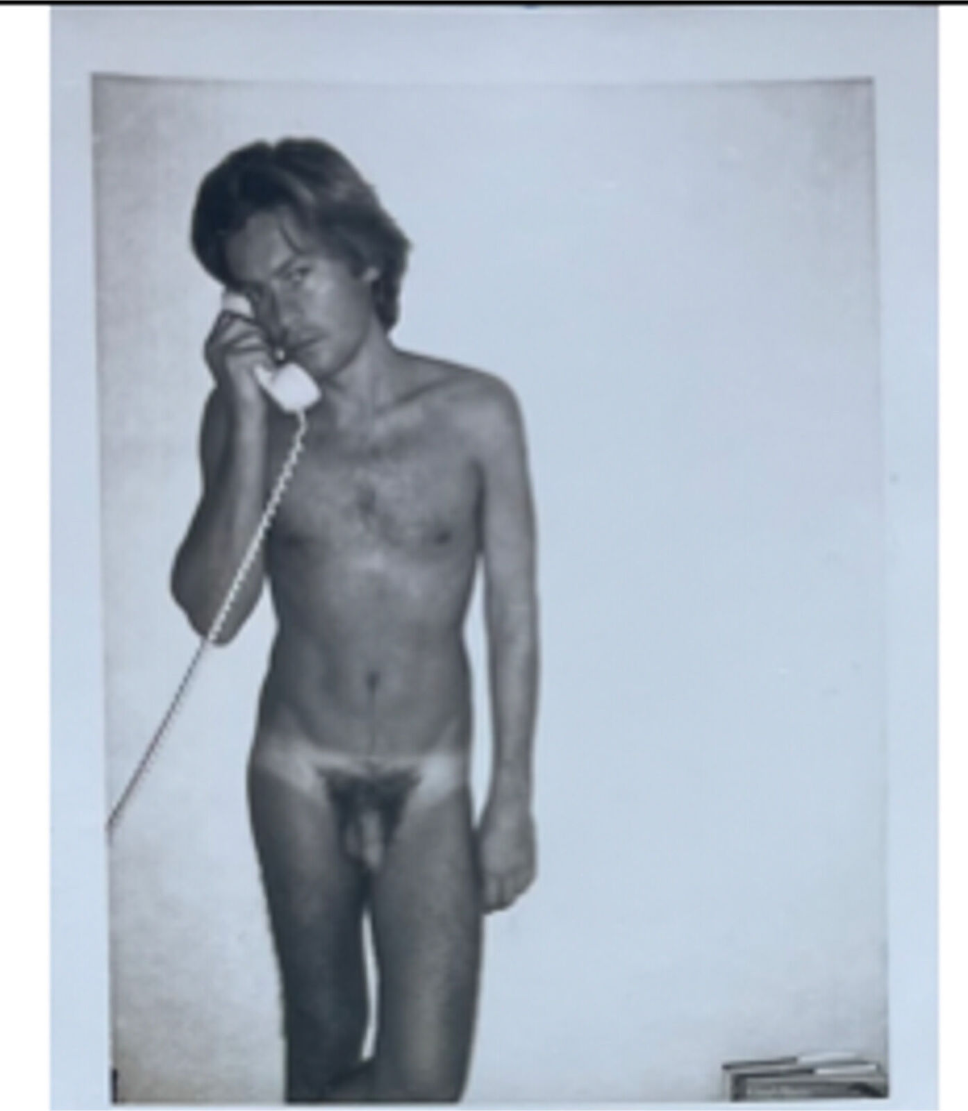 A naked man talking on a telephone while standing in front of a wall.