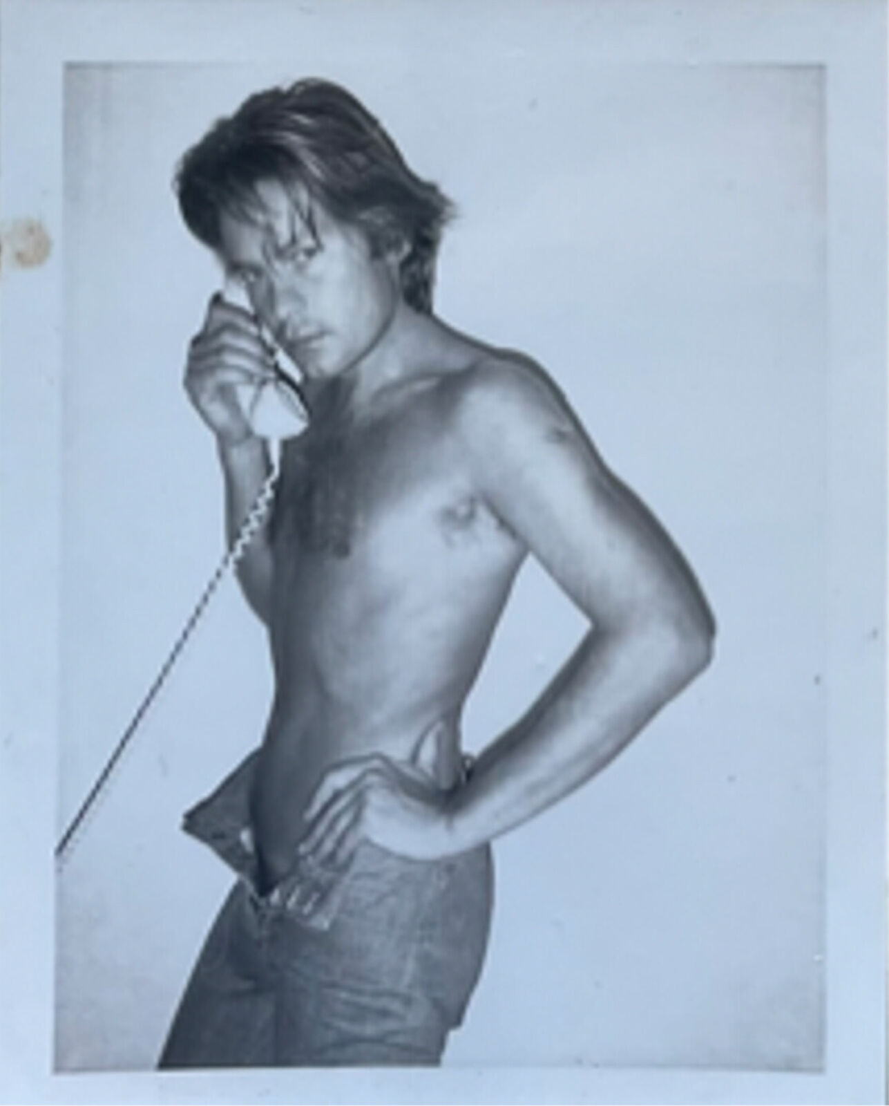 A shirtless man talking on a telephone while standing in front of a wall.