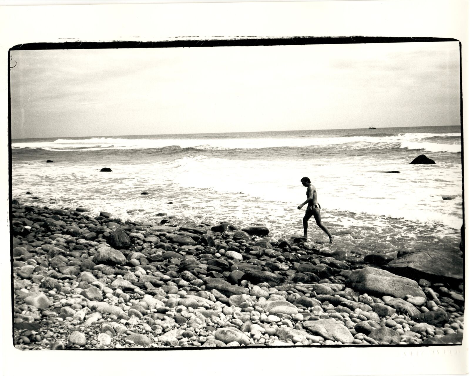 A black and white photo of a person walking on a rocky beach.