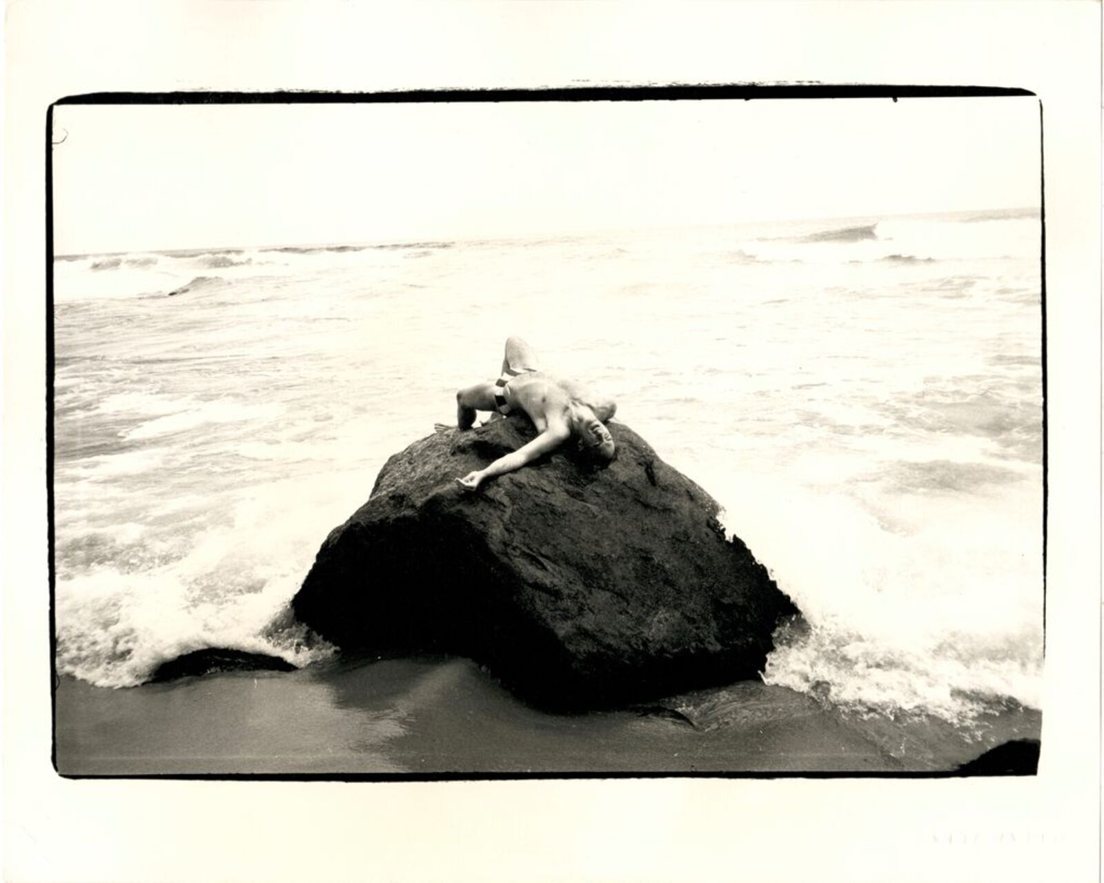 A black and white photo of a person laying on a rock in the ocean.