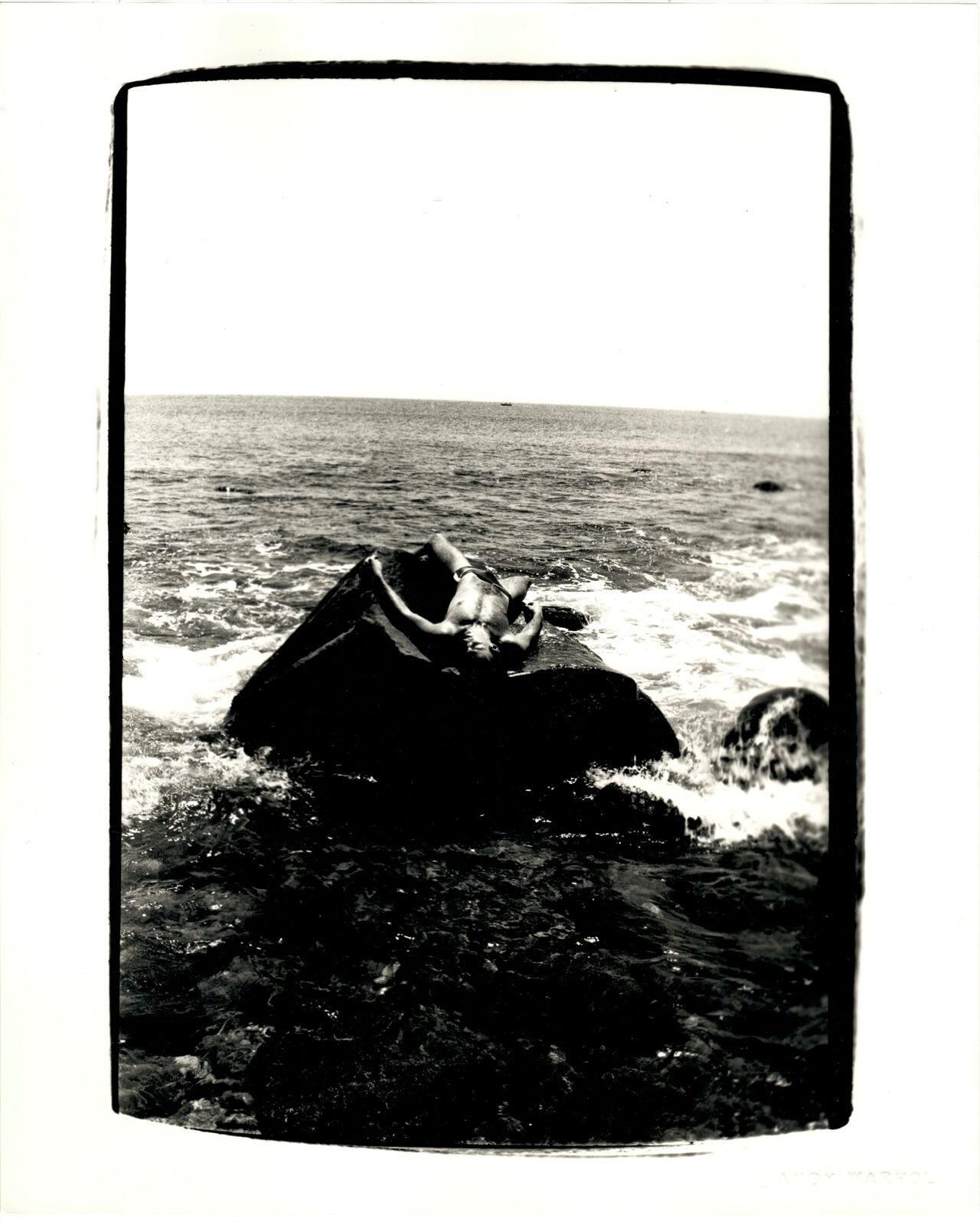 A black and white photo of a person laying on a rock in the ocean.