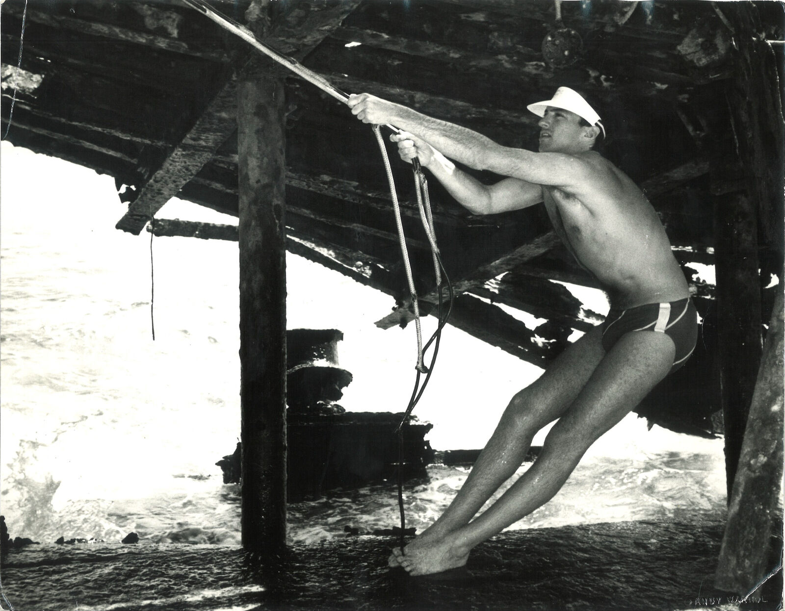 A man in a bathing suit and hat holding onto a rope.