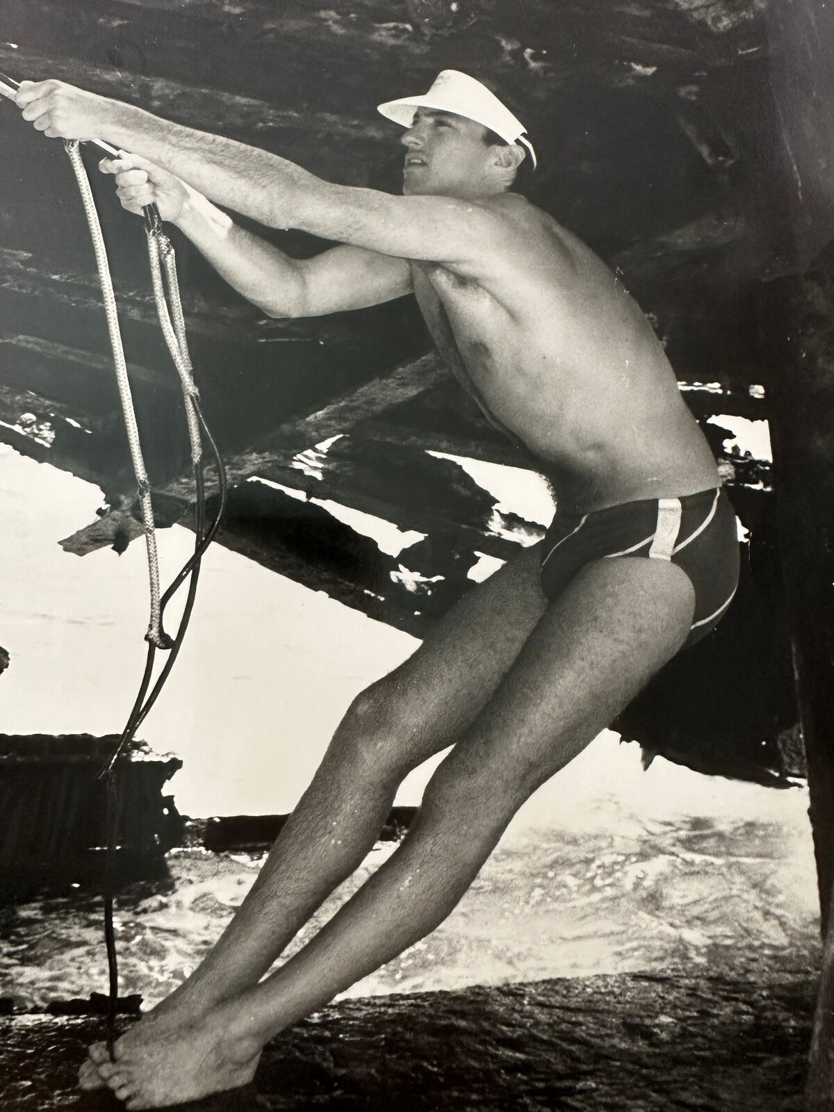 A man in a bathing suit and hat holding onto a rope.