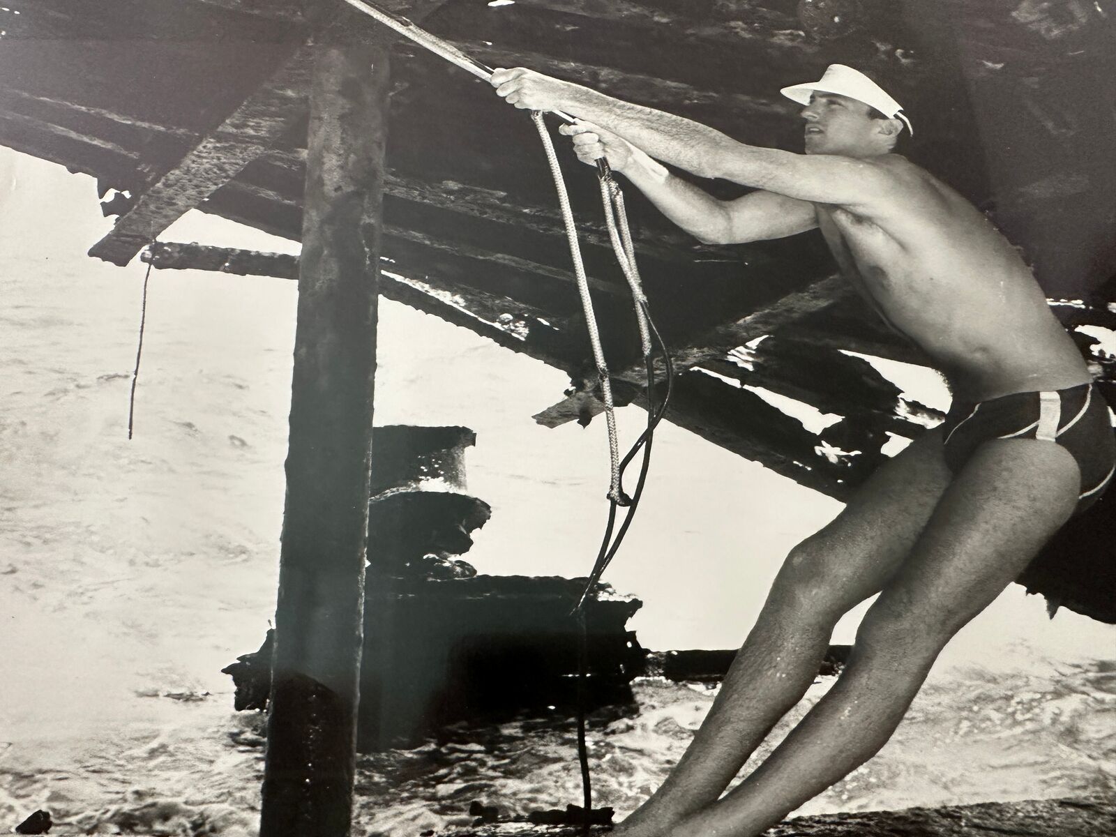 A man in a bathing suit and hat holding onto a rope.