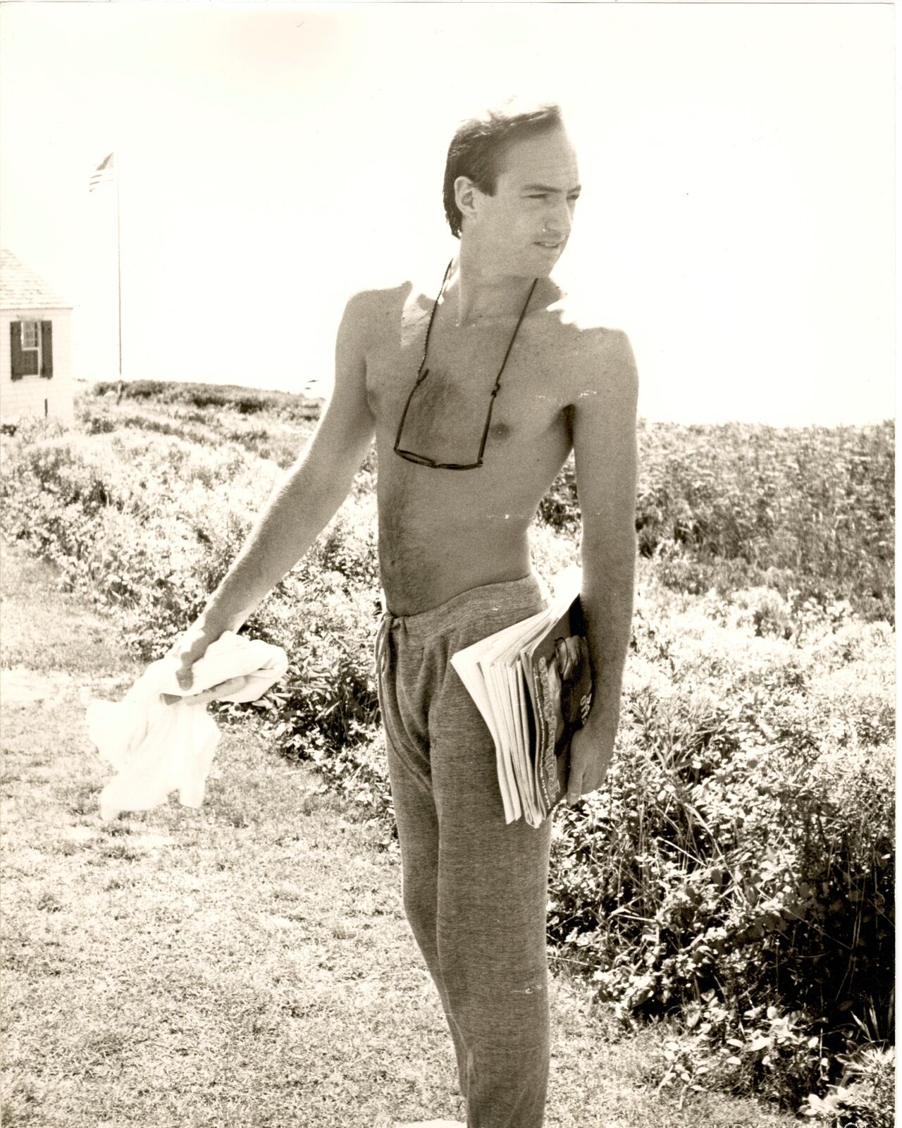 A shirtless man standing in a field holding a newspaper.