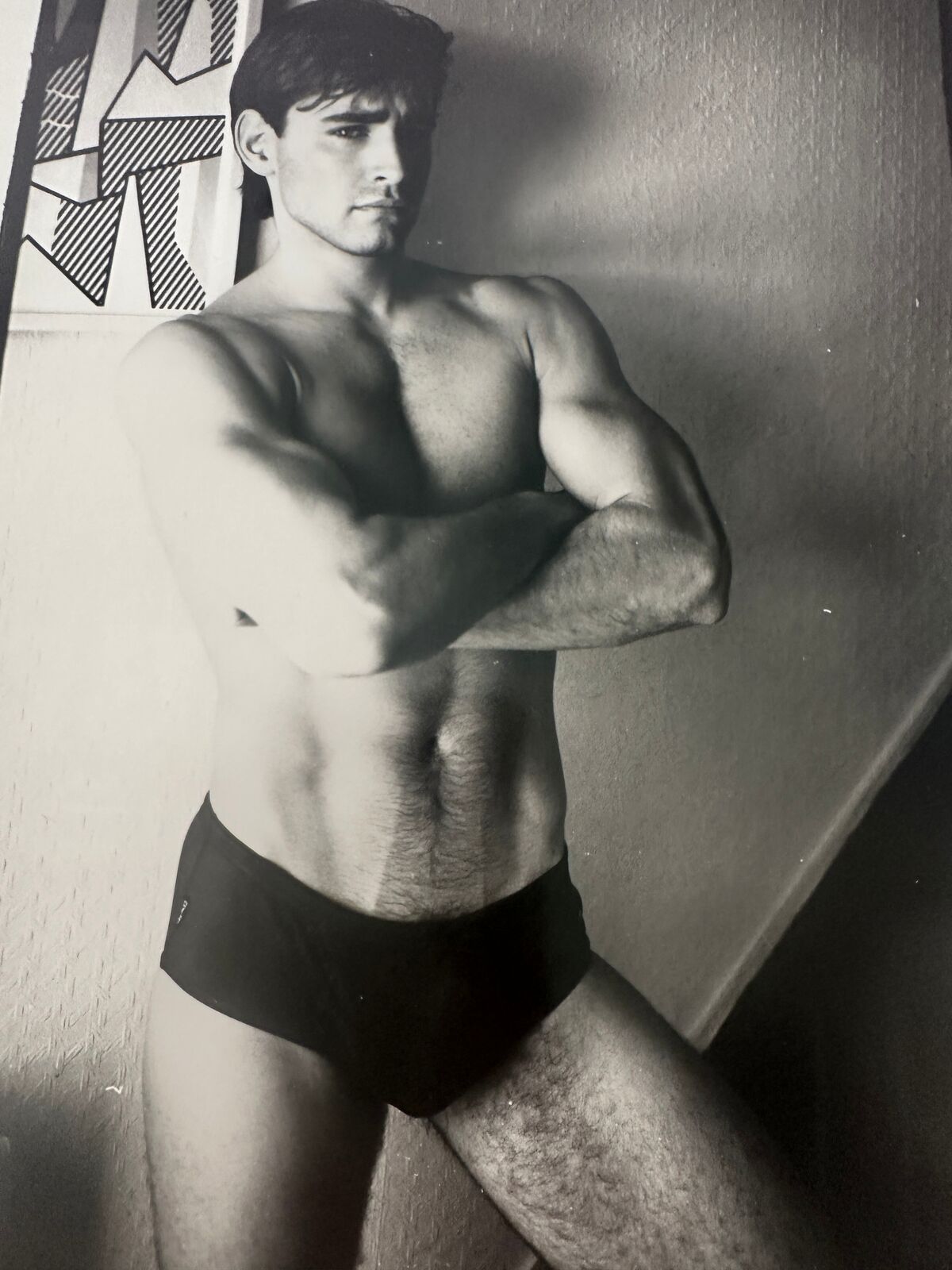 A shirtless man in a black underwear leaning against a wall.