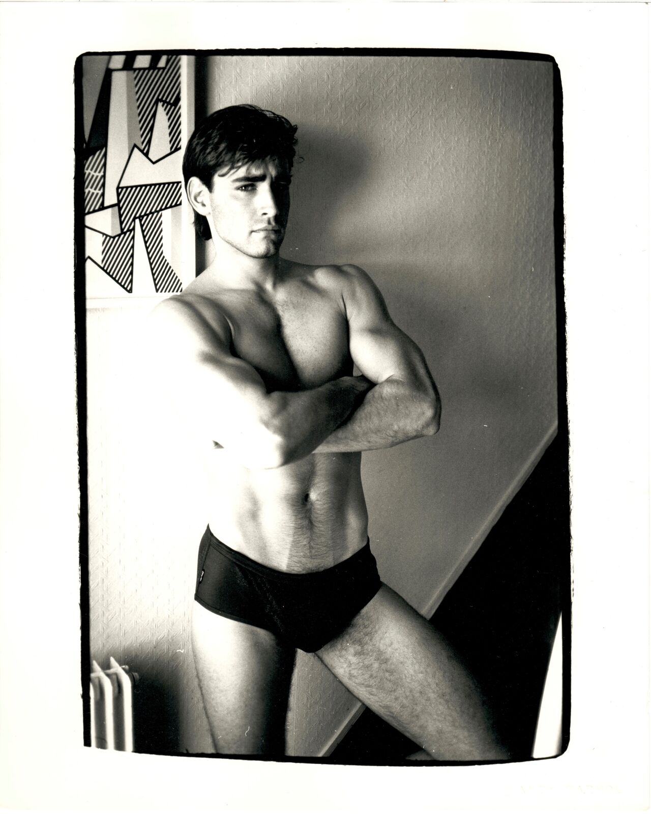 A shirtless man in a black underwear leaning against a wall.
