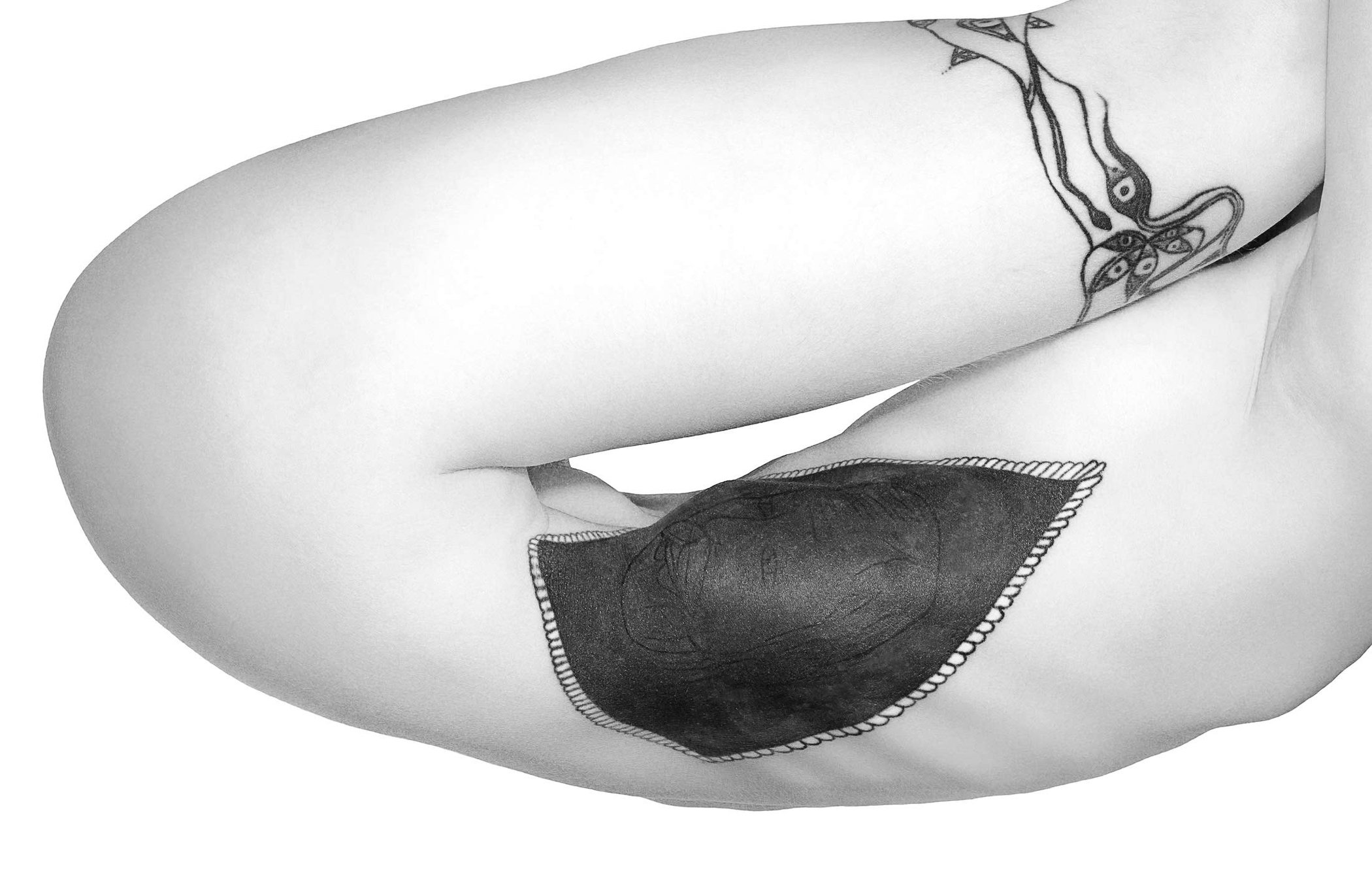 A black-and-white close-up of a person's arm featuring a dark, stitched tattoo resembling a leaf or shield.