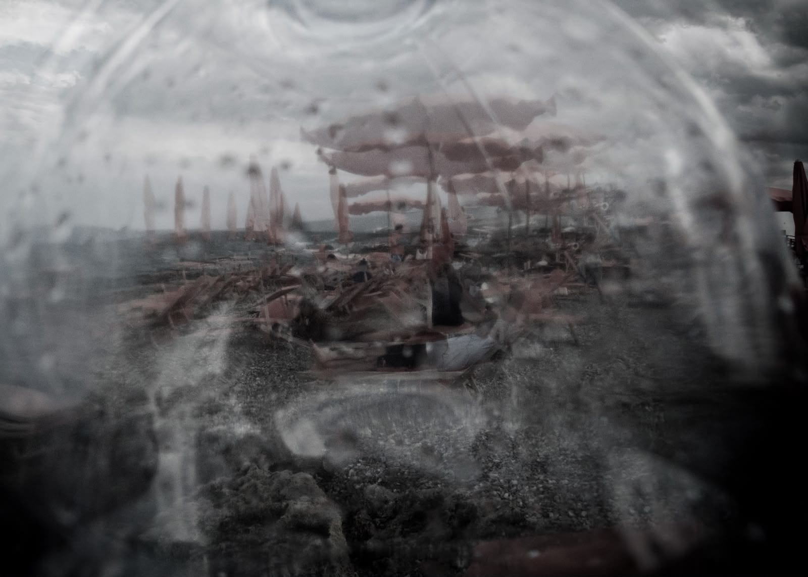 A view through a textured glass surface reveals a desolate beach scene with scattered lounge chairs and umbrellas under an overcast sky.