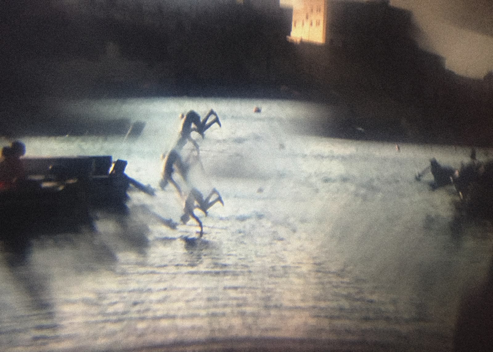 A grainy, low-light photograph captures a figure mid-air over water with a distant building in the background.