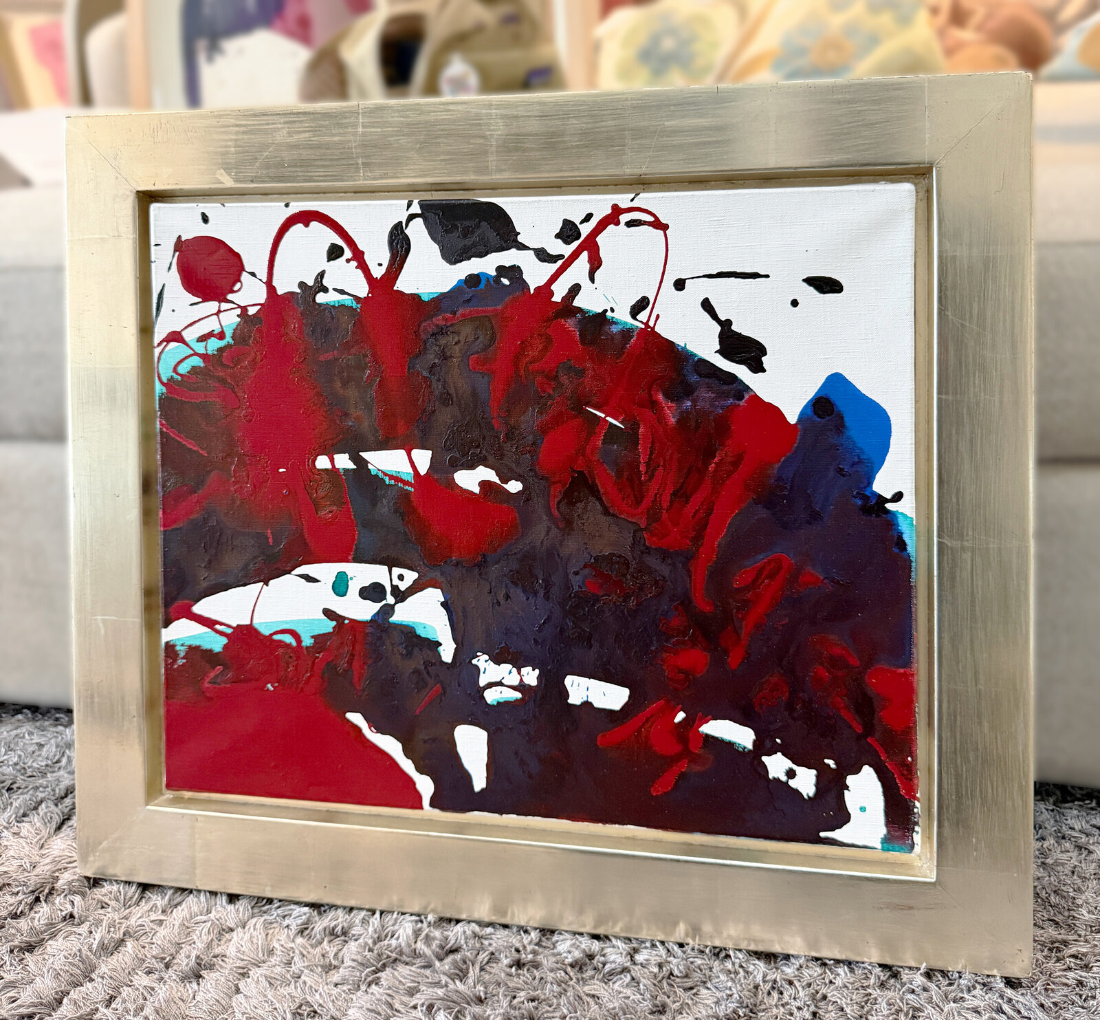 An abstract composition featuring bold red and black splatters against a white background, framed in metallic silver.