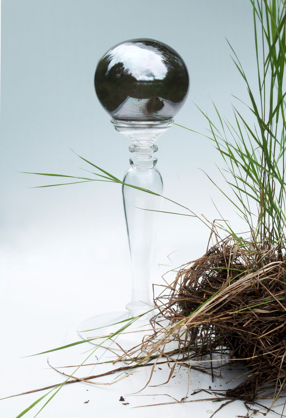 A clear glass candlestick with a reflective sphere top stands beside a bundle of dried grass against a white background.