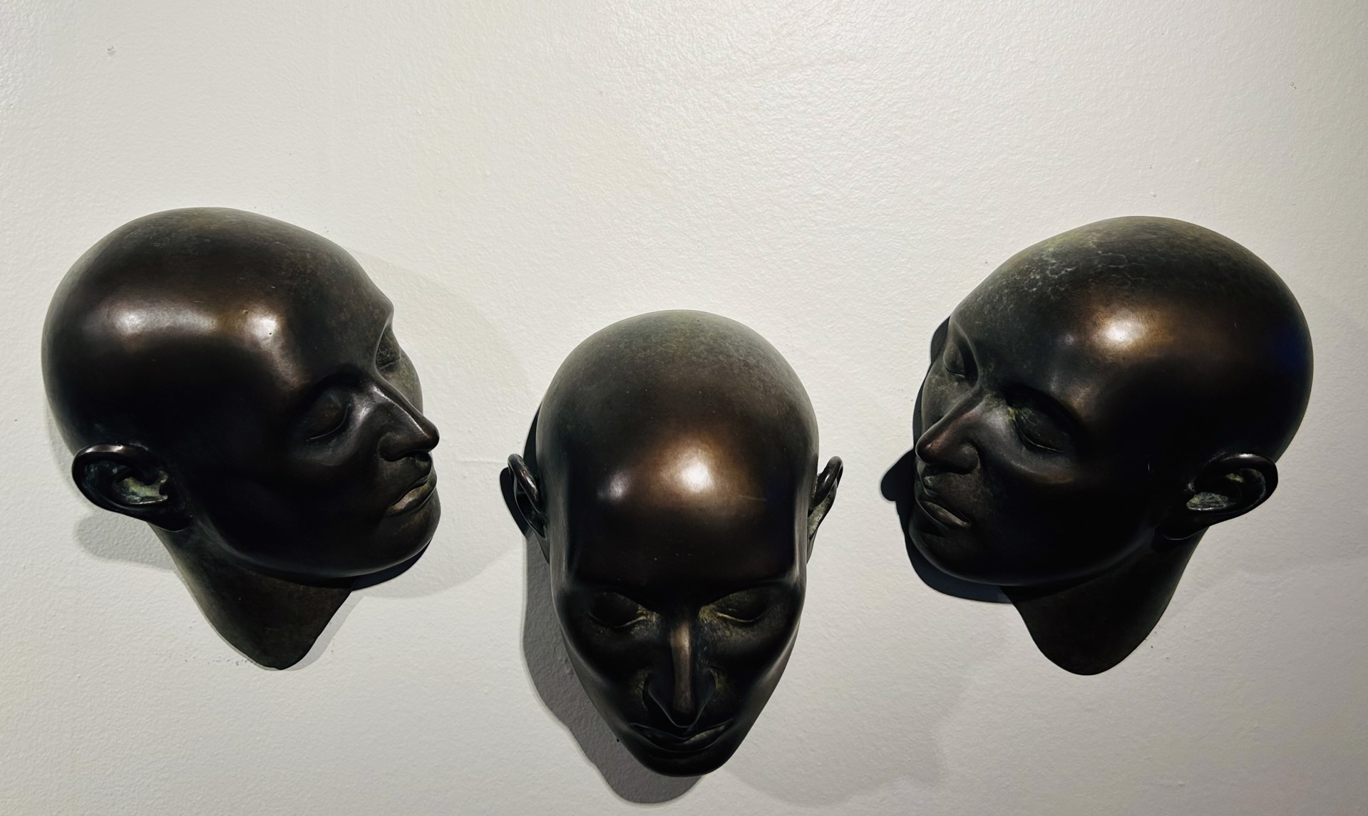 Three dark bronze mannequin heads mounted on a plain wall in a minimalist arrangement.