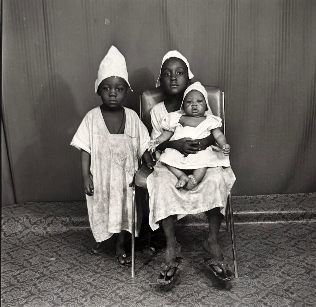A black-and-white portrait of three young figures in matching white head coverings and loose garments, posed formally against a patterned backdrop.