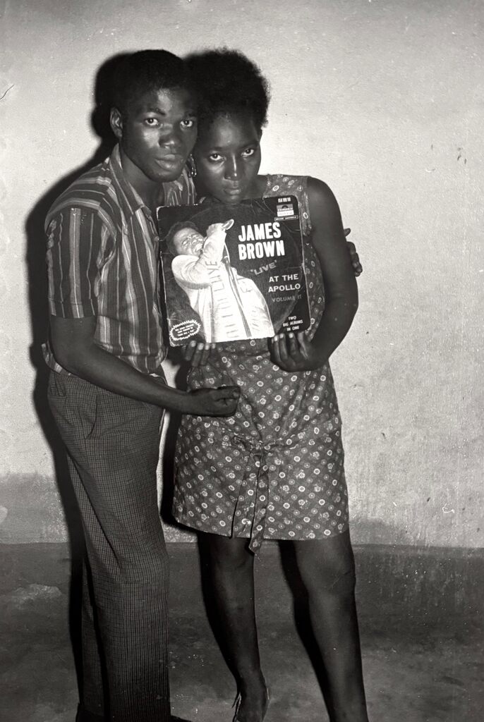 A black-and-white portrait of two figures standing close together, holding an album cover.