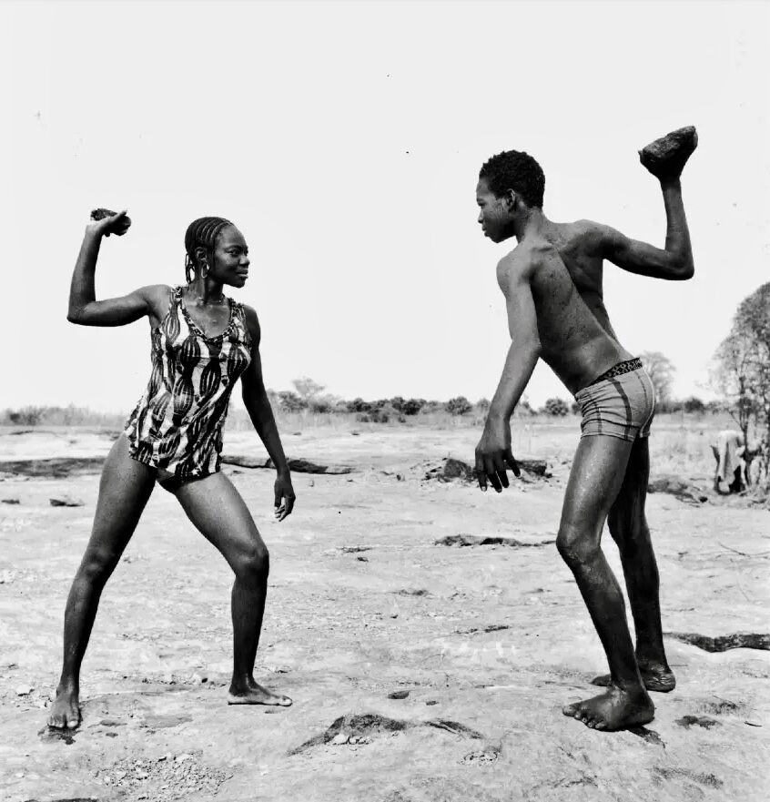 A black-and-white photograph captures two figures in a dynamic, confrontational pose against an open landscape.