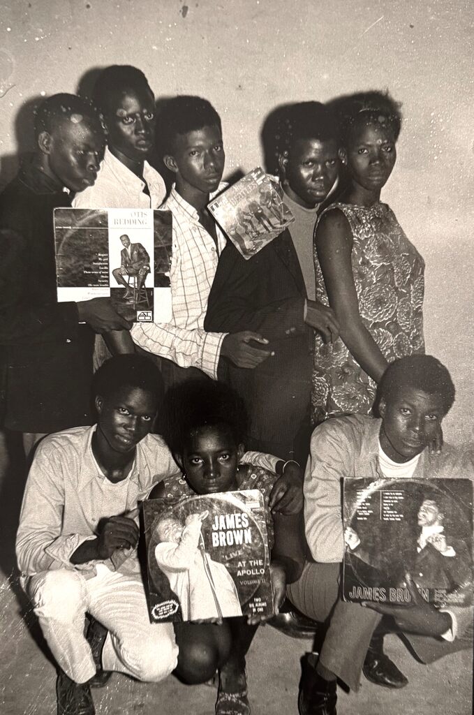 A black-and-white group portrait of seven individuals holding vinyl records against a plain wall.