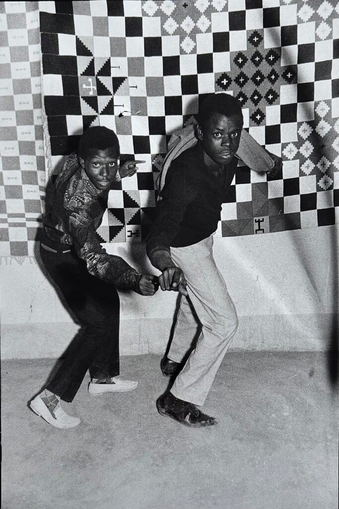 Two figures in a dynamic pose against a geometric quilted backdrop.