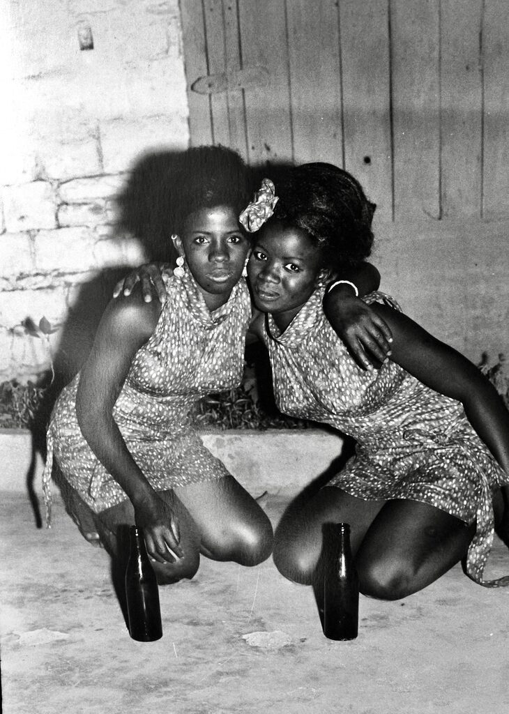 A black-and-white photograph of two figures in patterned dresses crouching together with bottles on the ground.