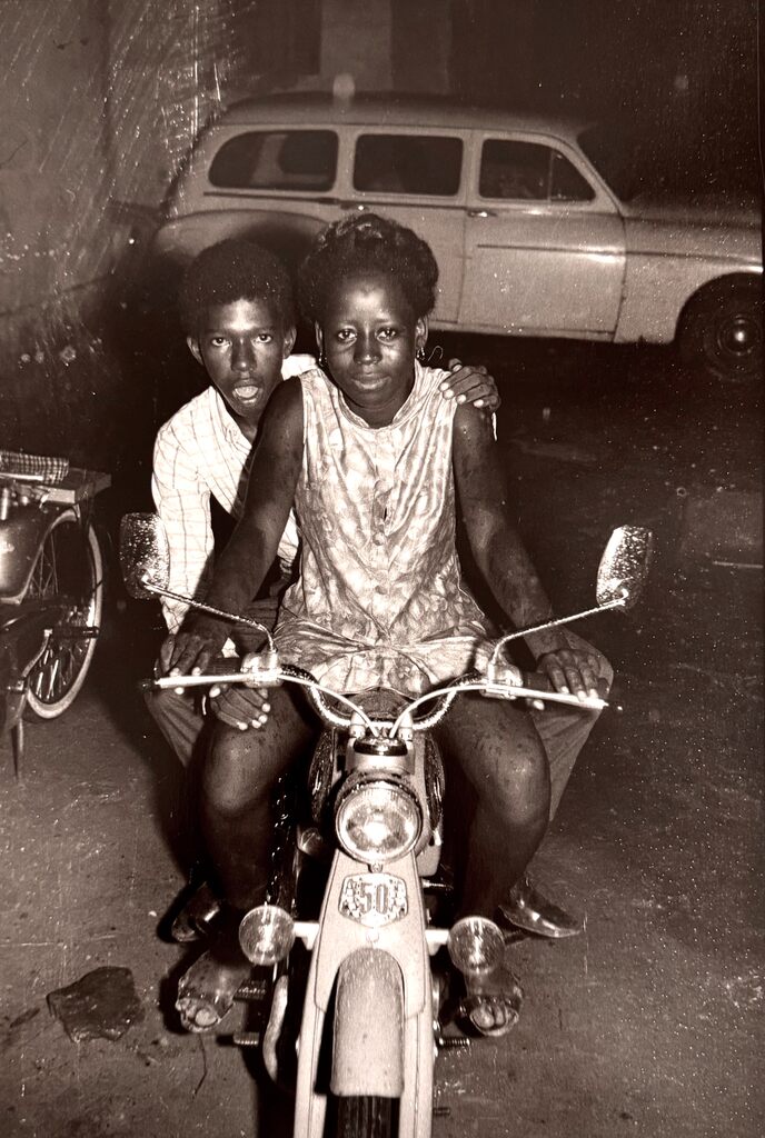 A black-and-white photograph captures two figures on a motorcycle, with a vintage car in the background.