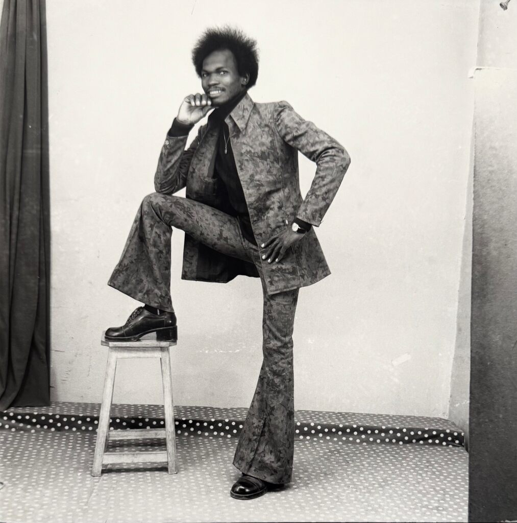 A stylish figure in a patterned suit poses confidently on a stool against a plain backdrop.