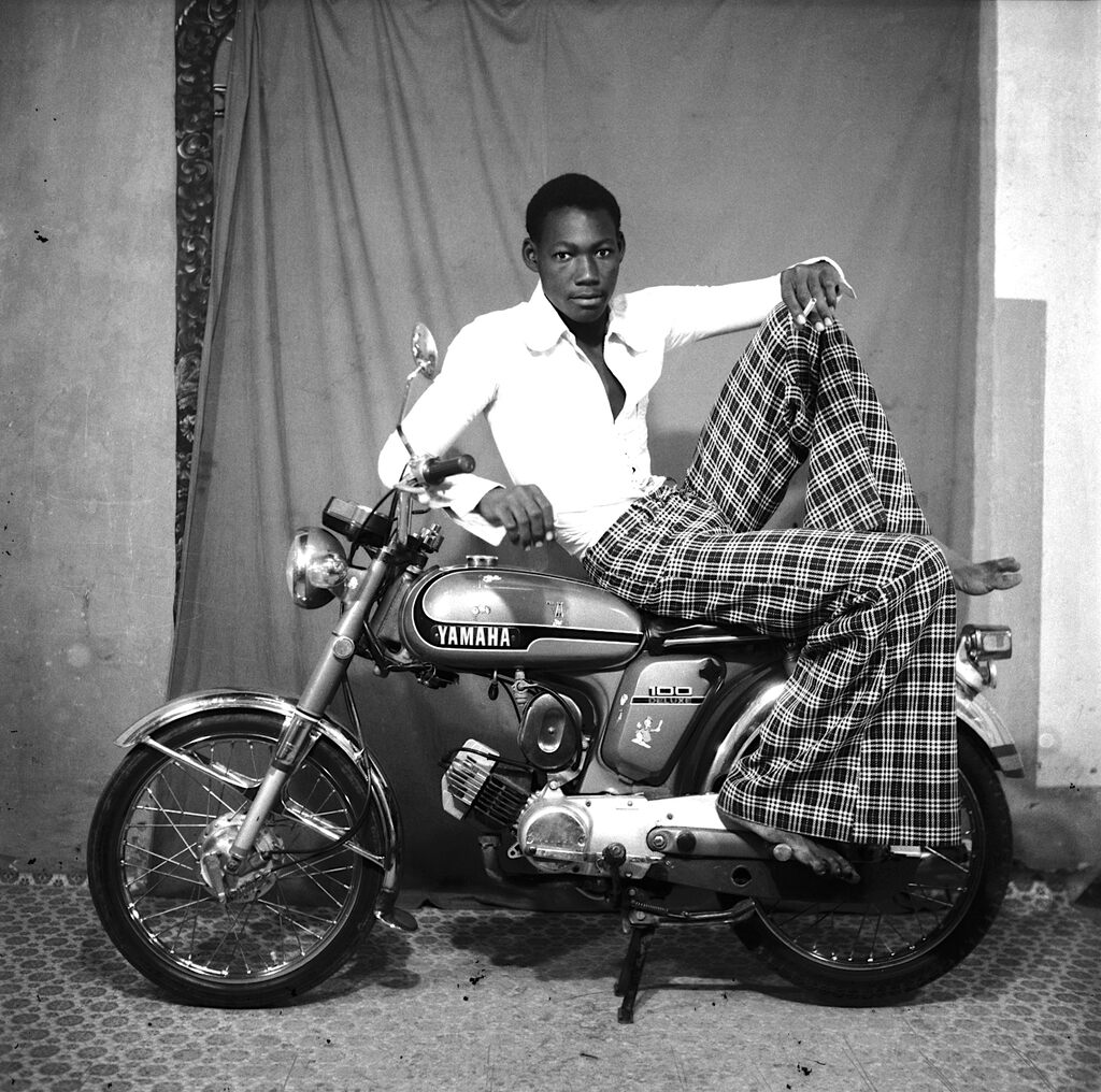 A relaxed figure sits casually on a vintage motorbike in a studio setting.