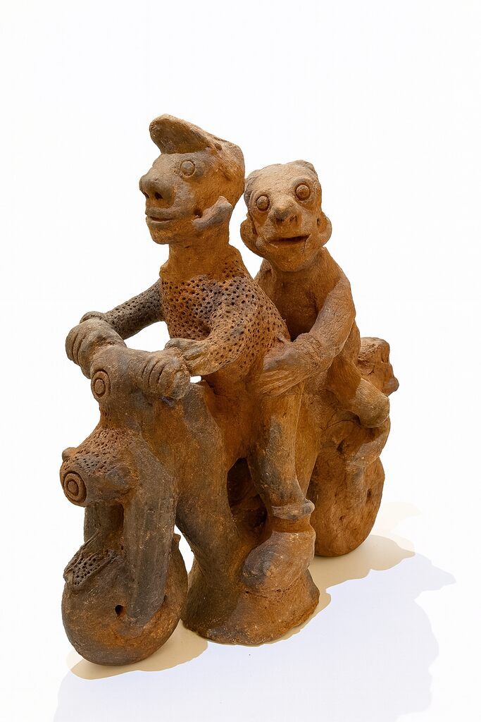 A rustic terracotta sculpture of two figures riding a motorcycle, featuring textured surfaces and whimsical expressions.