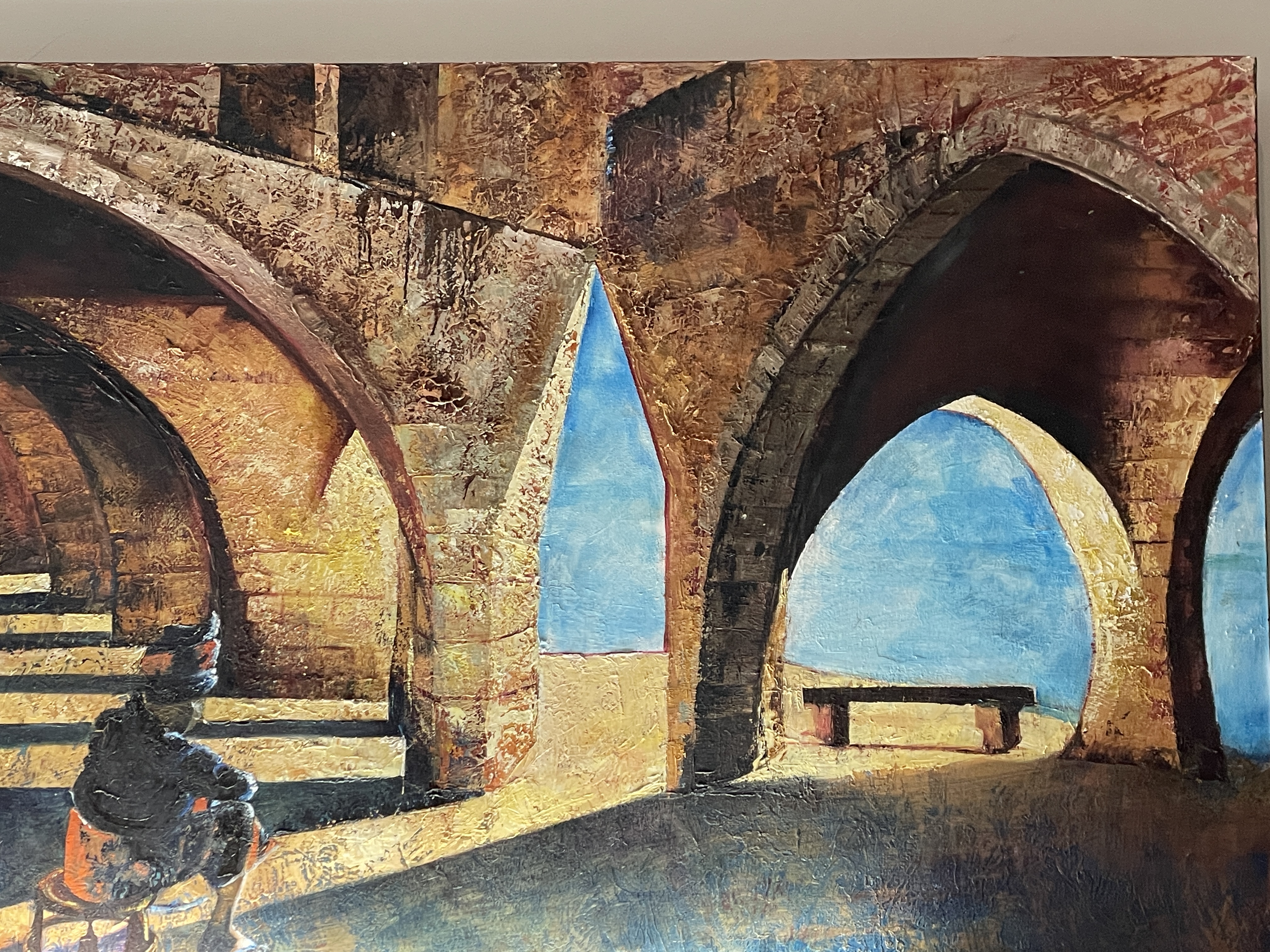 A gallery space features a large painting of an arched stone structure with a seated figure, displayed above a black bench.