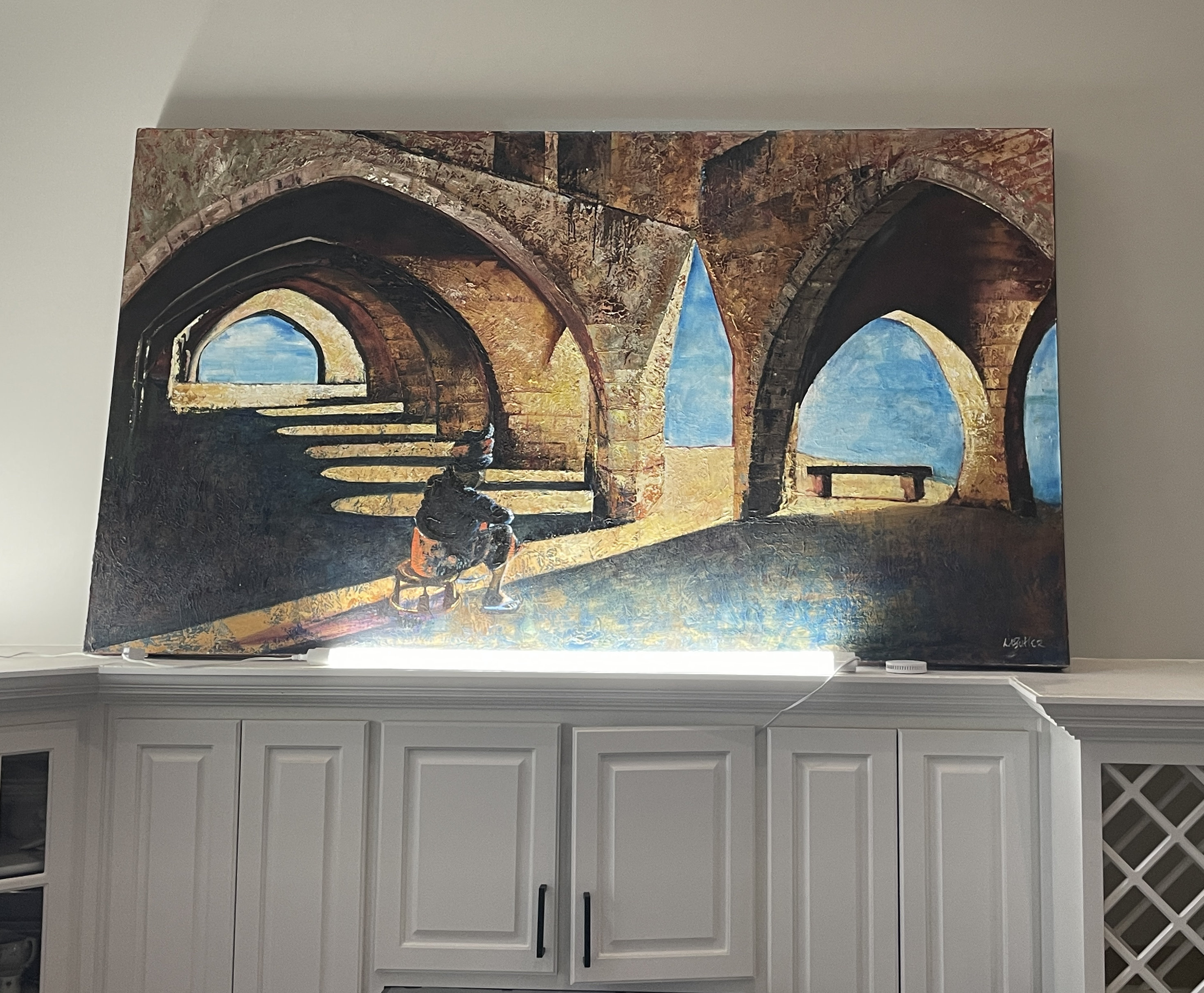 A gallery space features a large painting of an arched stone structure with a seated figure, displayed above a black bench.