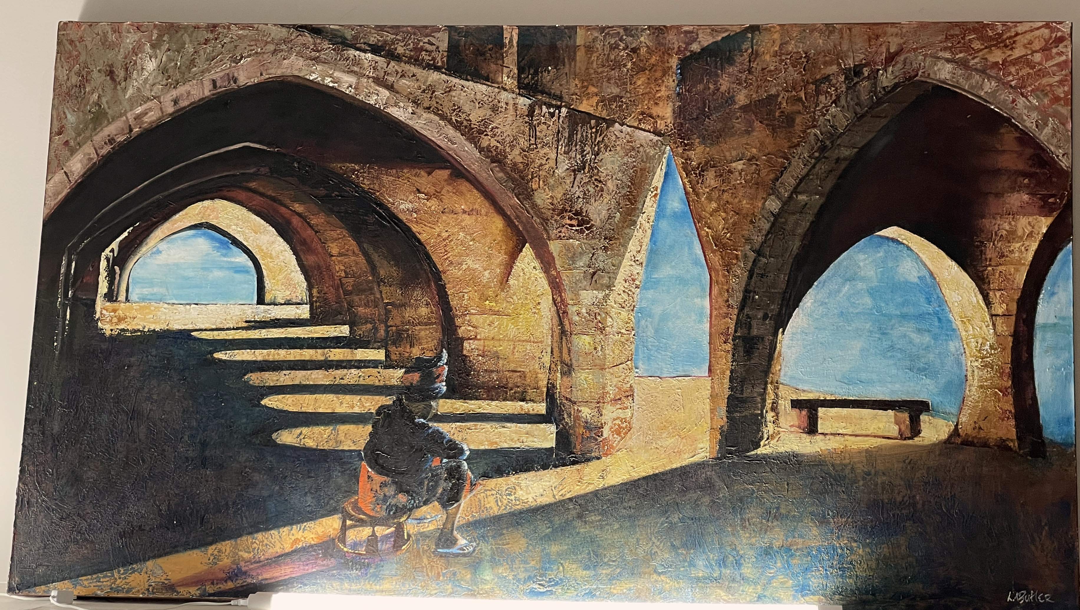 A gallery space features a large painting of an arched stone structure with a seated figure, displayed above a black bench.