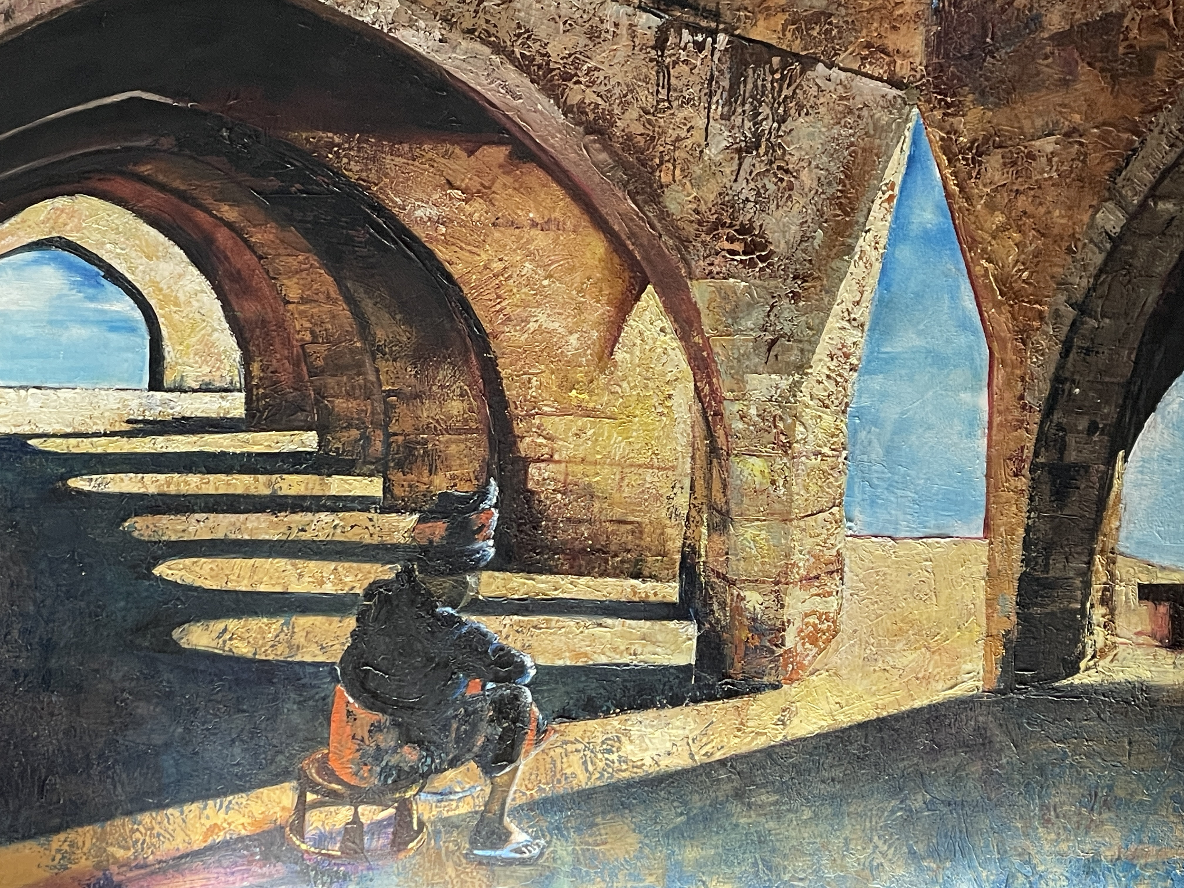 A gallery space features a large painting of an arched stone structure with a seated figure, displayed above a black bench.