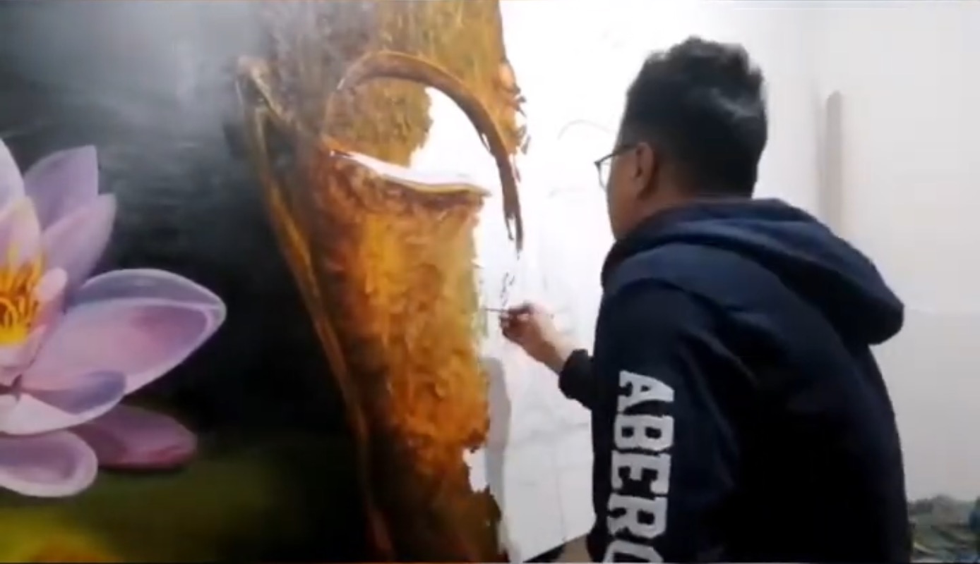 A person stands in a gallery, observing a large painting of a golden face flanked by pink lotus flowers.
