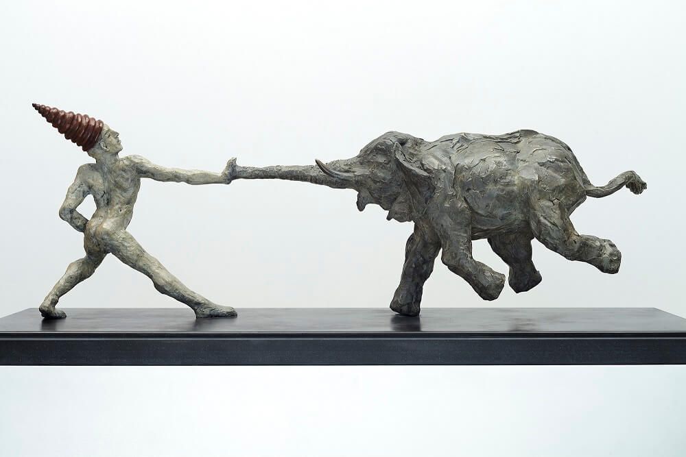 A dynamic bronze sculpture of a figure pulling an elephant by the trunk.