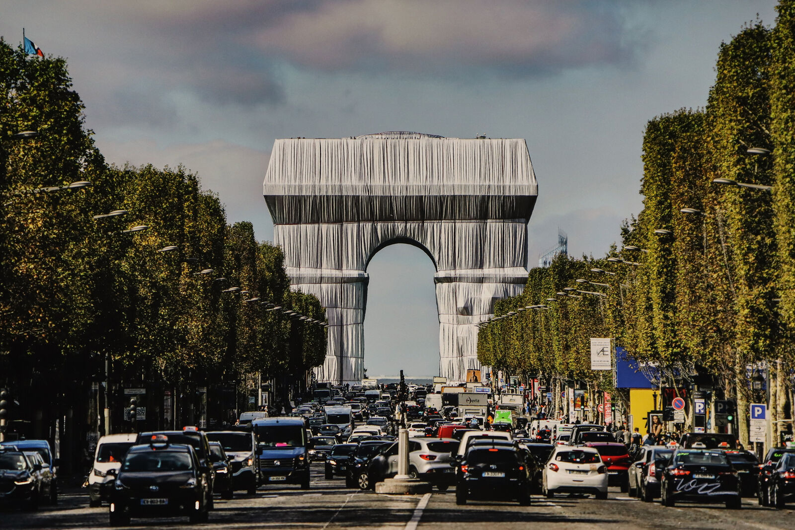 A wide avenue filled with traffic leads to a massive arch draped in white fabric.