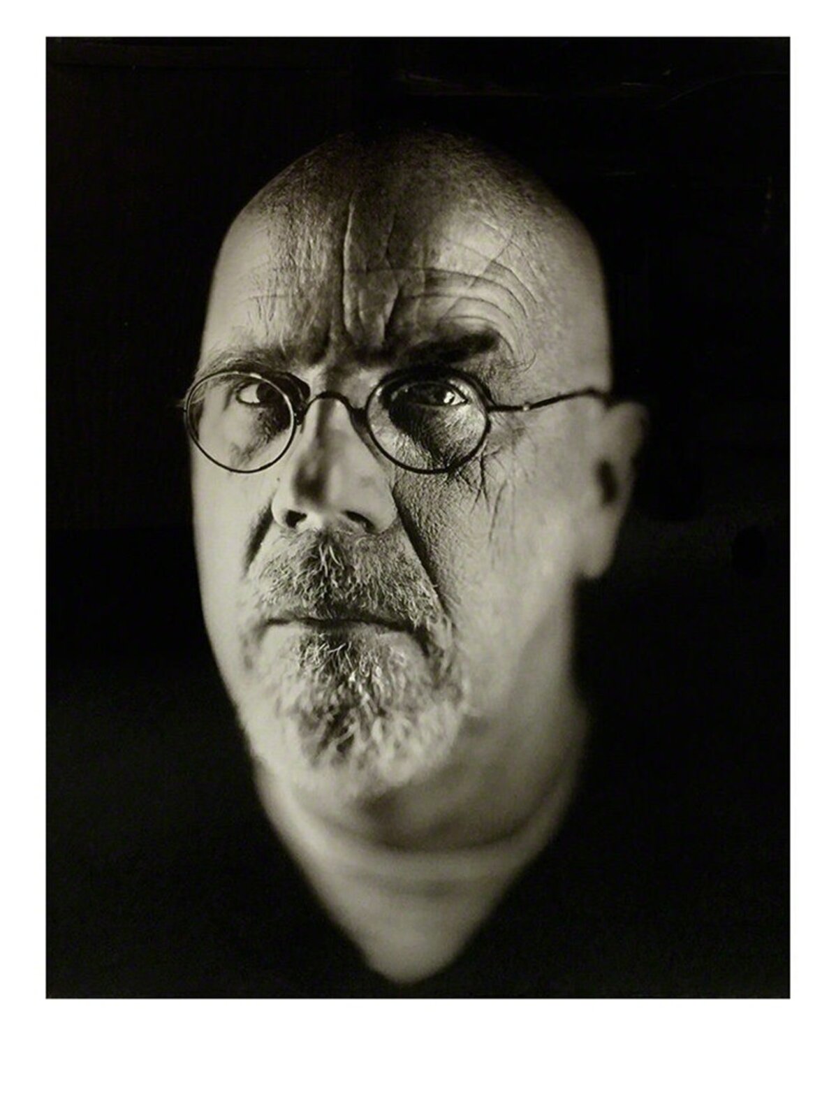 A monochrome portrait of a bald figure with glasses and a goatee, captured in dramatic lighting.