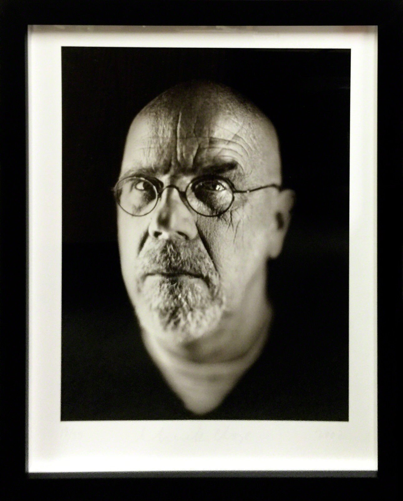 A monochrome portrait of a bald figure with glasses and a goatee, captured in dramatic lighting.